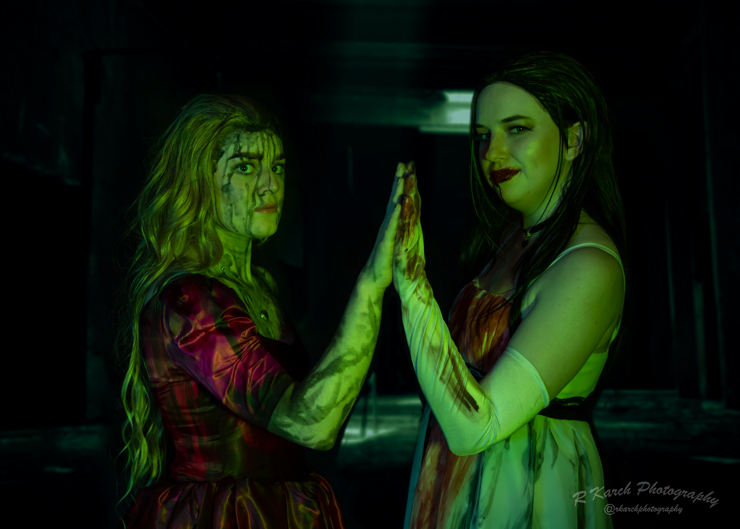 Two women in vintage dresses pose dramatically under eerie green lighting for a gothic horror photoshoot.
