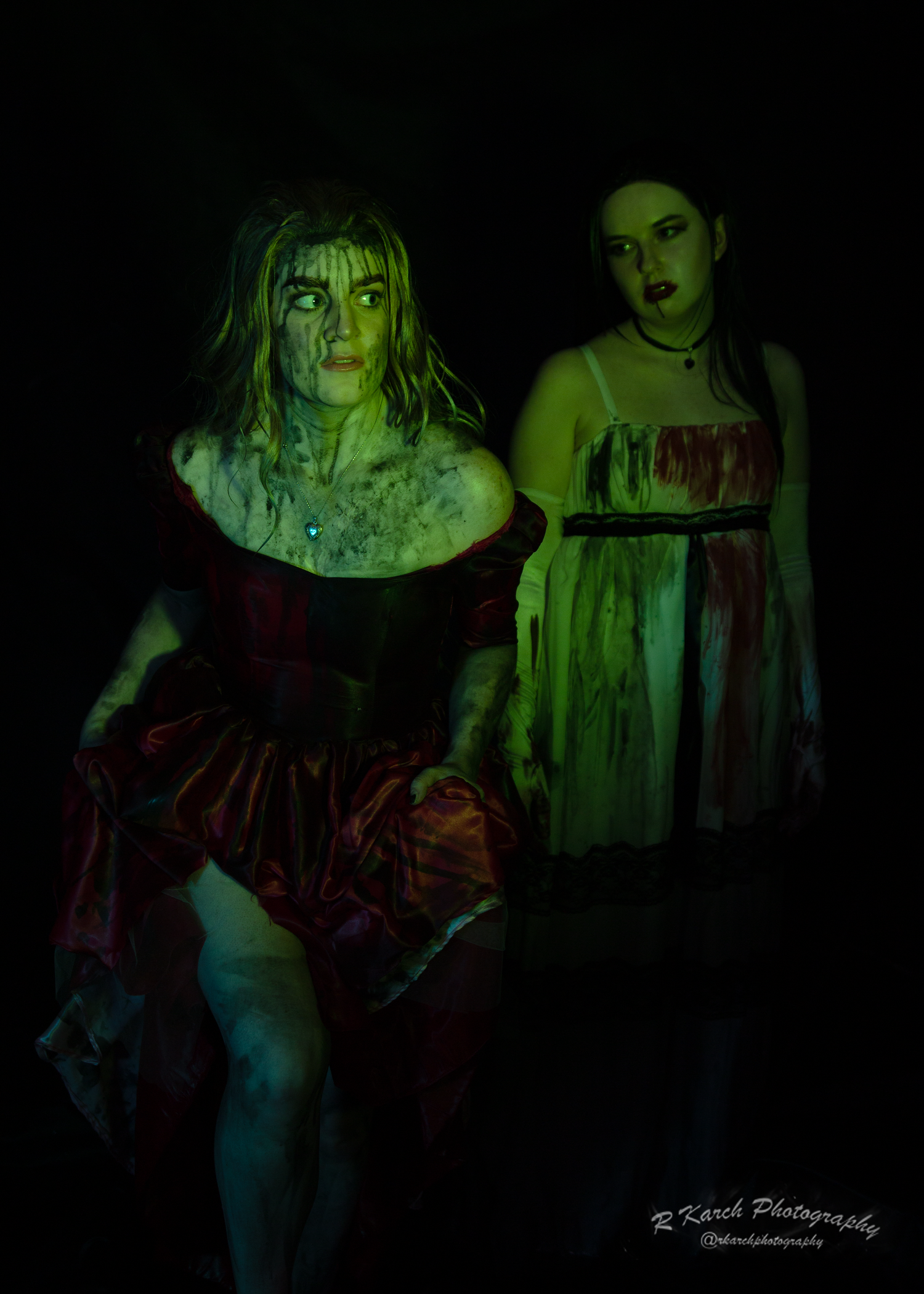 Two figures in blood-stained dresses stand menacingly in dark shadows with green lighting effects.