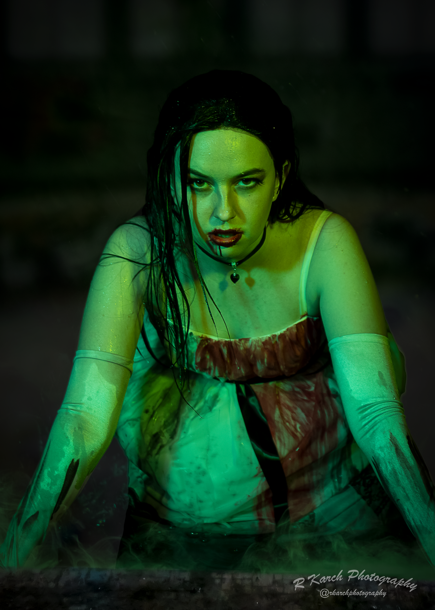 Gothic horror scene features performer in bloodstained dress crawling under green lighting.