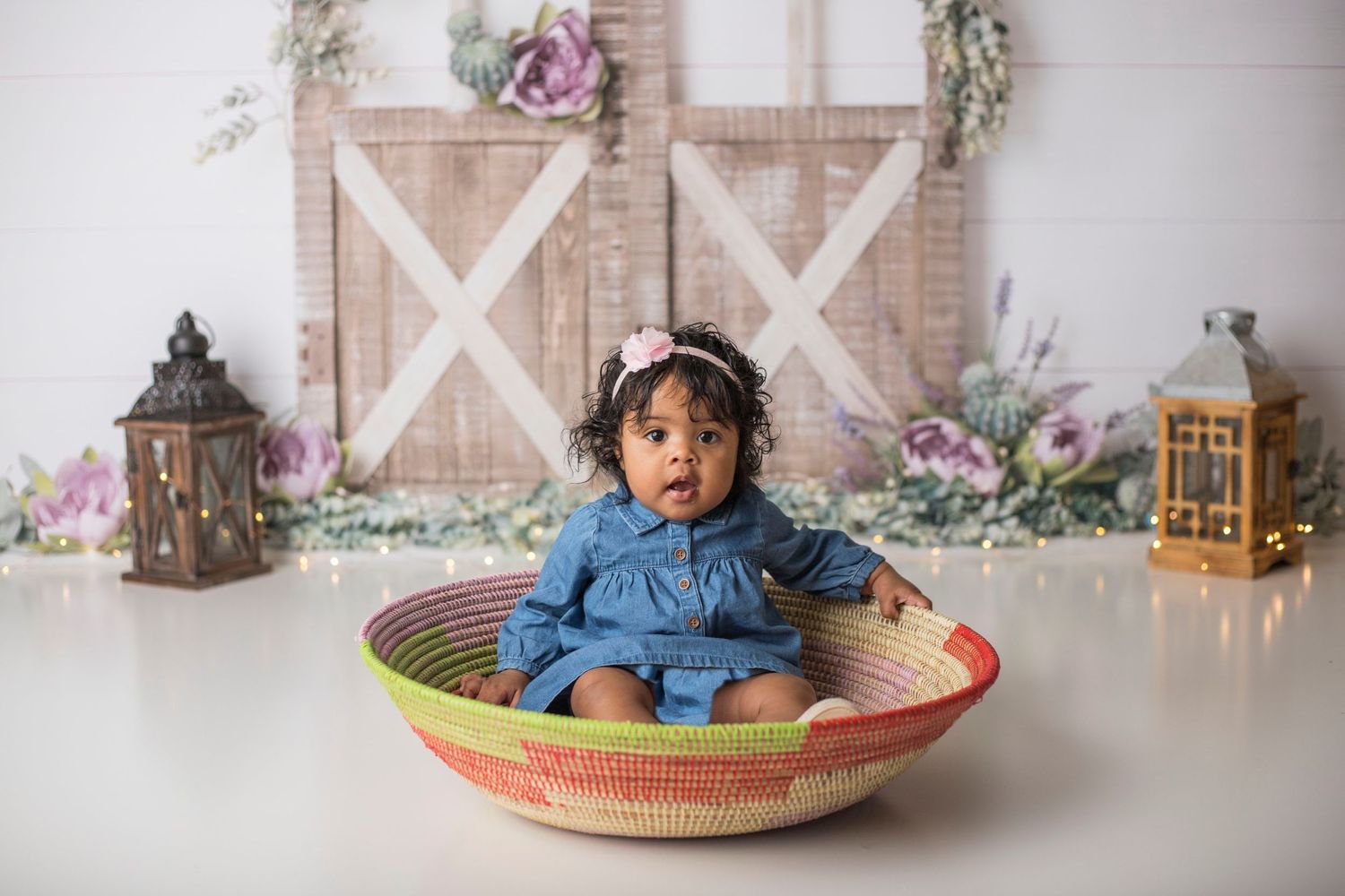 Baby in blue dress sits in woven basket against floral rustic backdrop with lanterns.