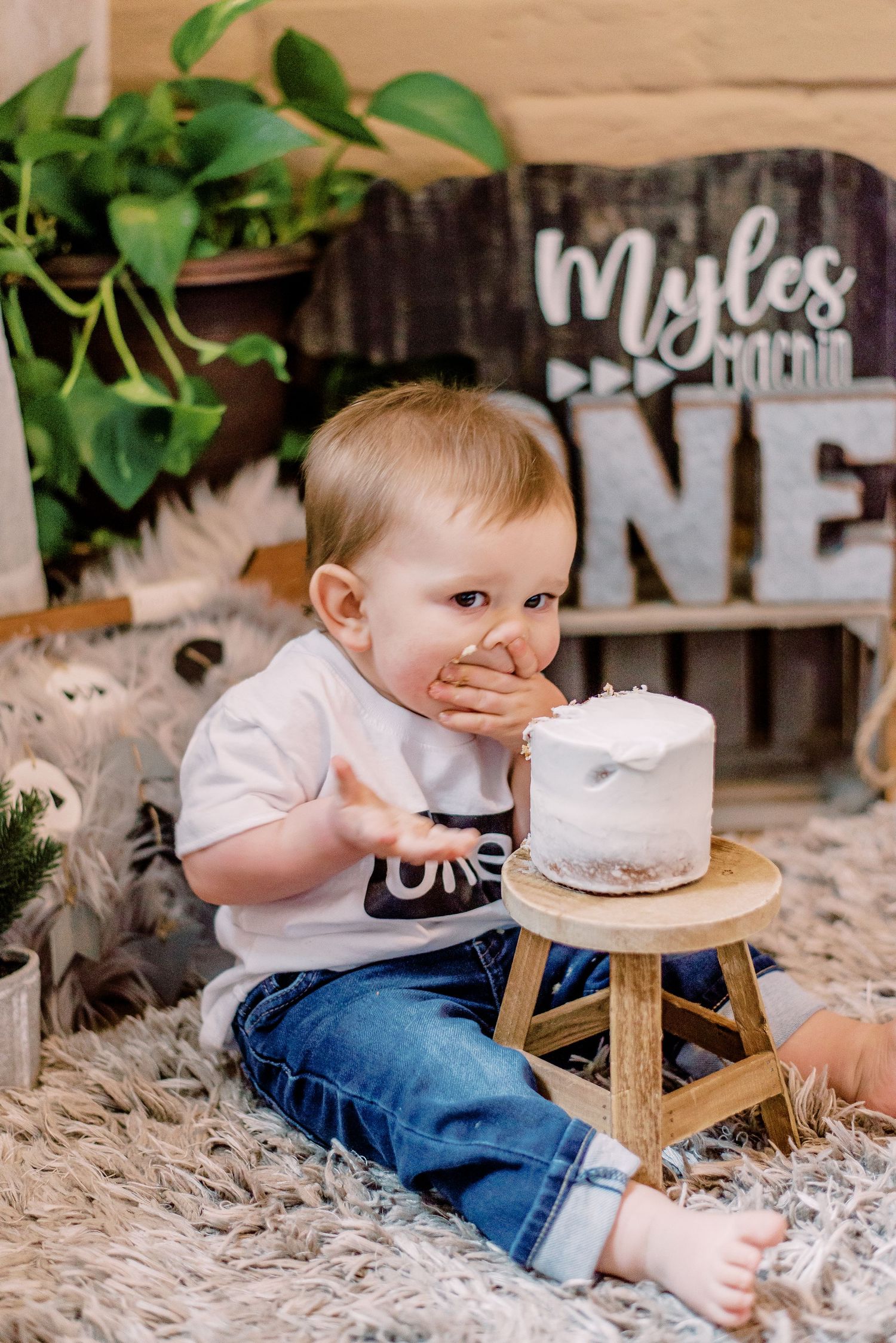 Baby in jeans and white shirt sits on fluffy rug with a small smash cake on wooden stool for first birthday celebration.