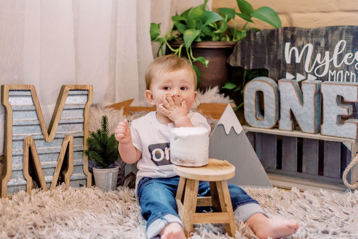 One year old birthday setup with wooden marquee letters, plants and rustic decor on cream carpet.