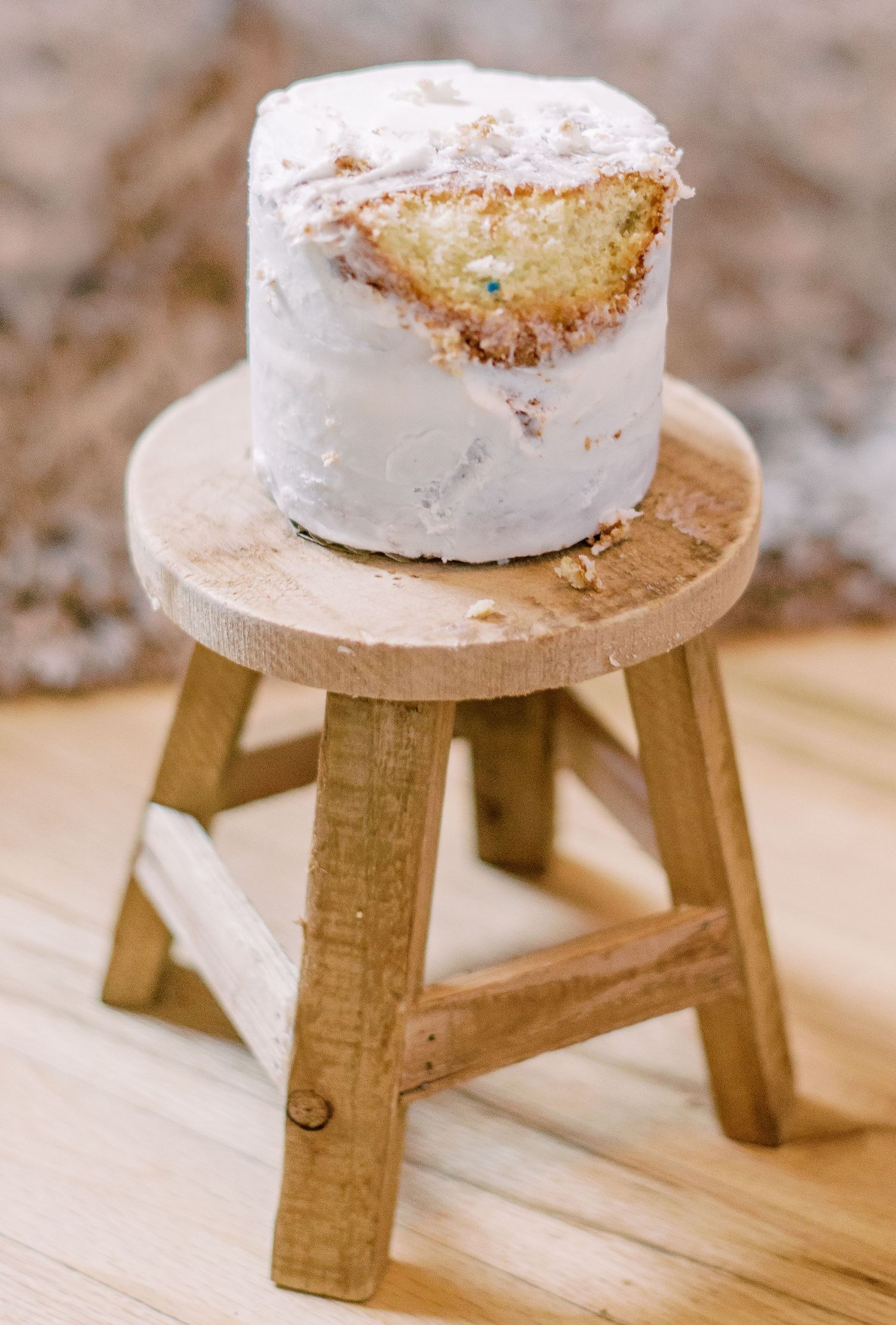 A white frosted cake slice sits atop a small wooden stool against a blurred beige background.
