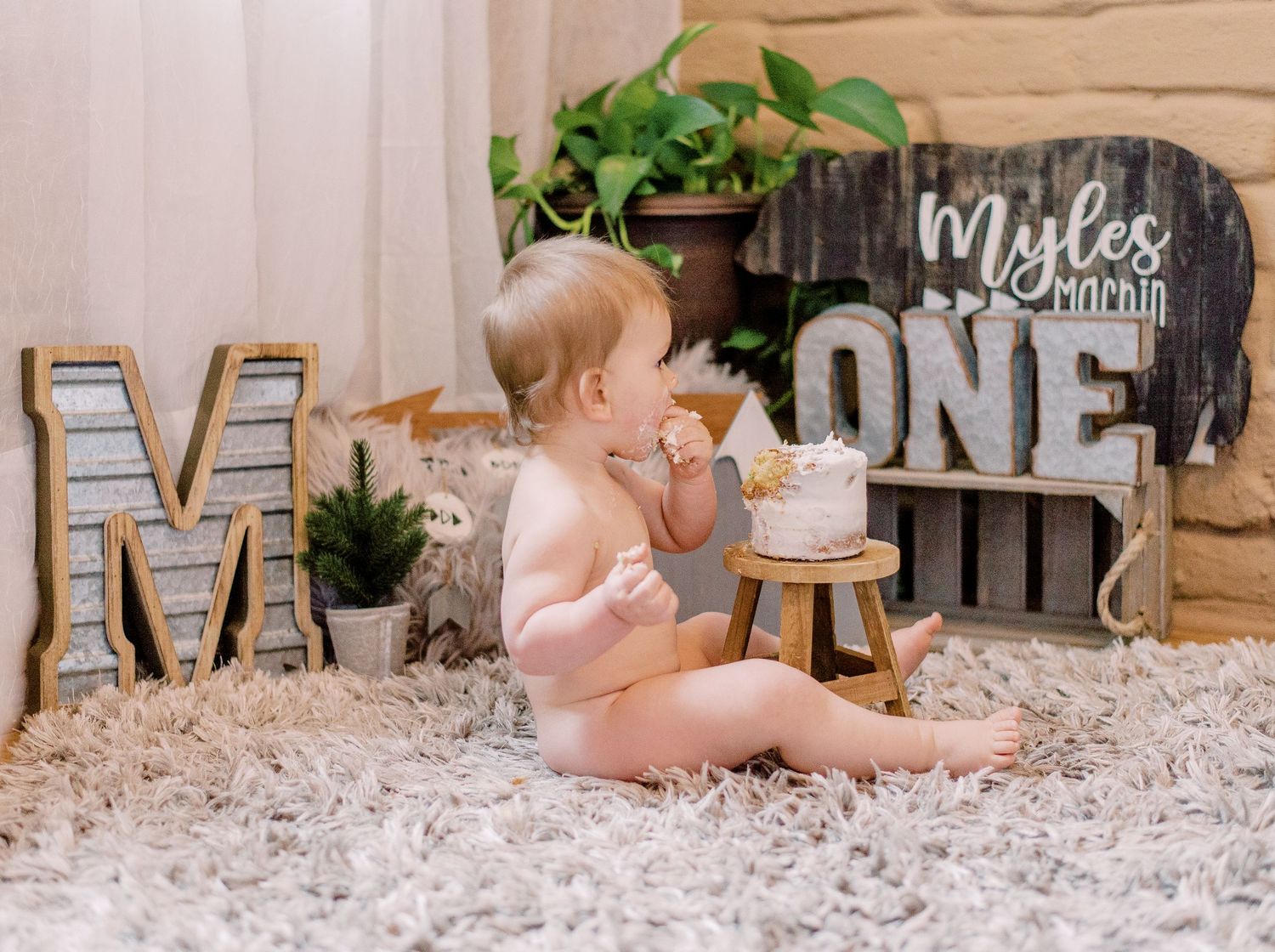 Baby in pink outfit sits on plush rug during first birthday cake smash photoshoot with rustic woodland decor.