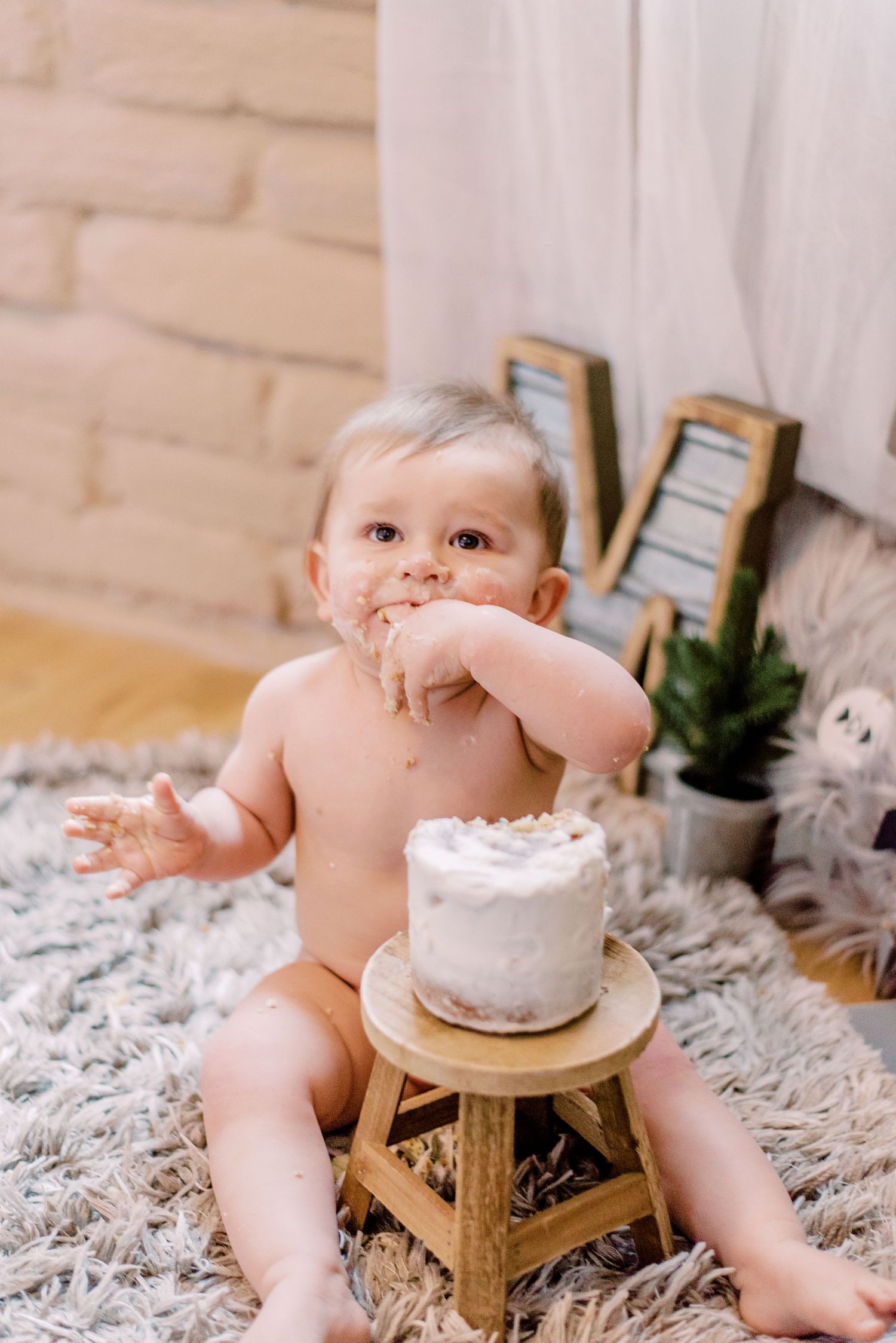 Baby in diaper sits on shag rug with small white birthday cake on wooden stool beside decorative plants.