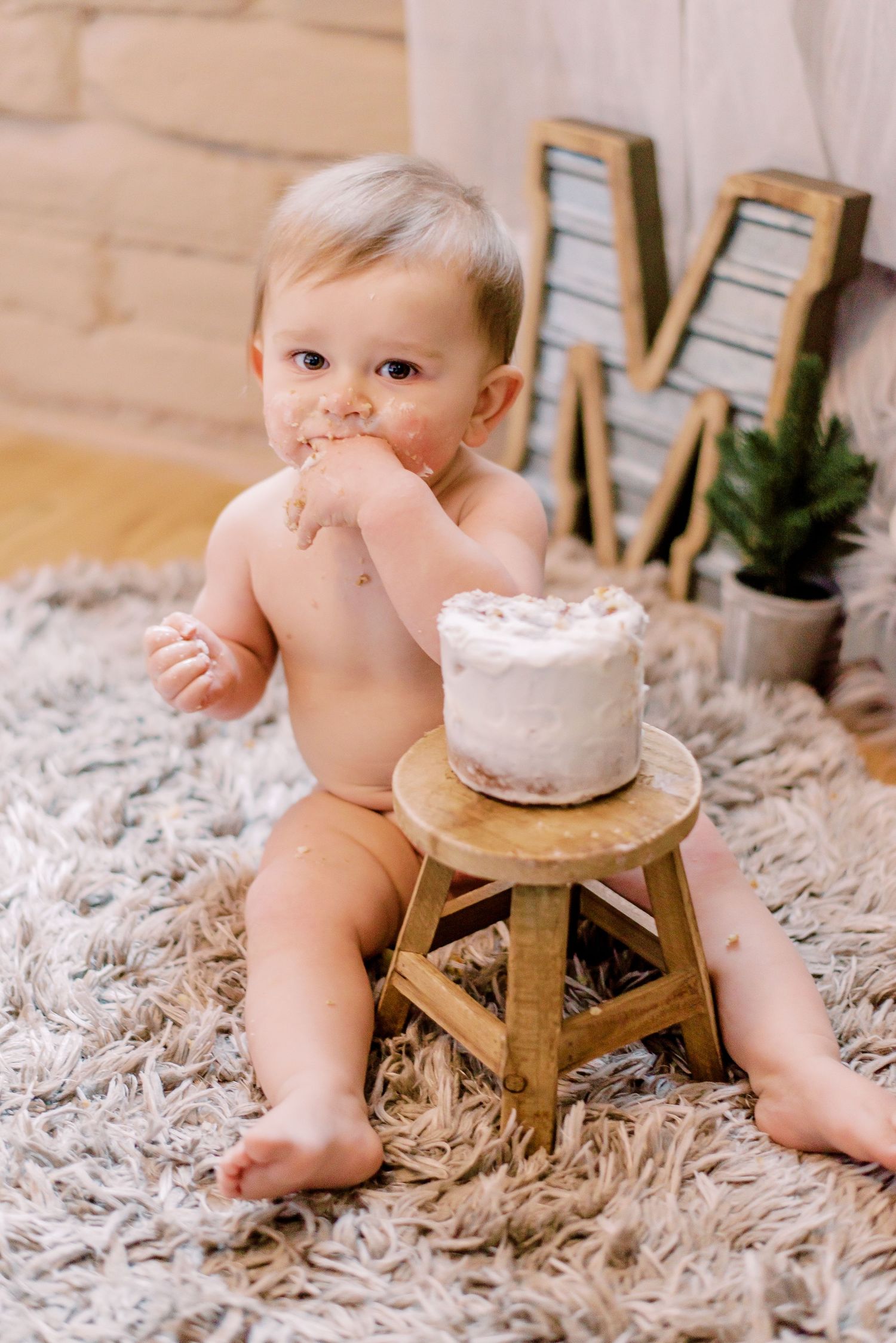 Small baby in diaper sitting on fluffy rug with white frosted cake on wooden stool and decorative letter M on wall.
