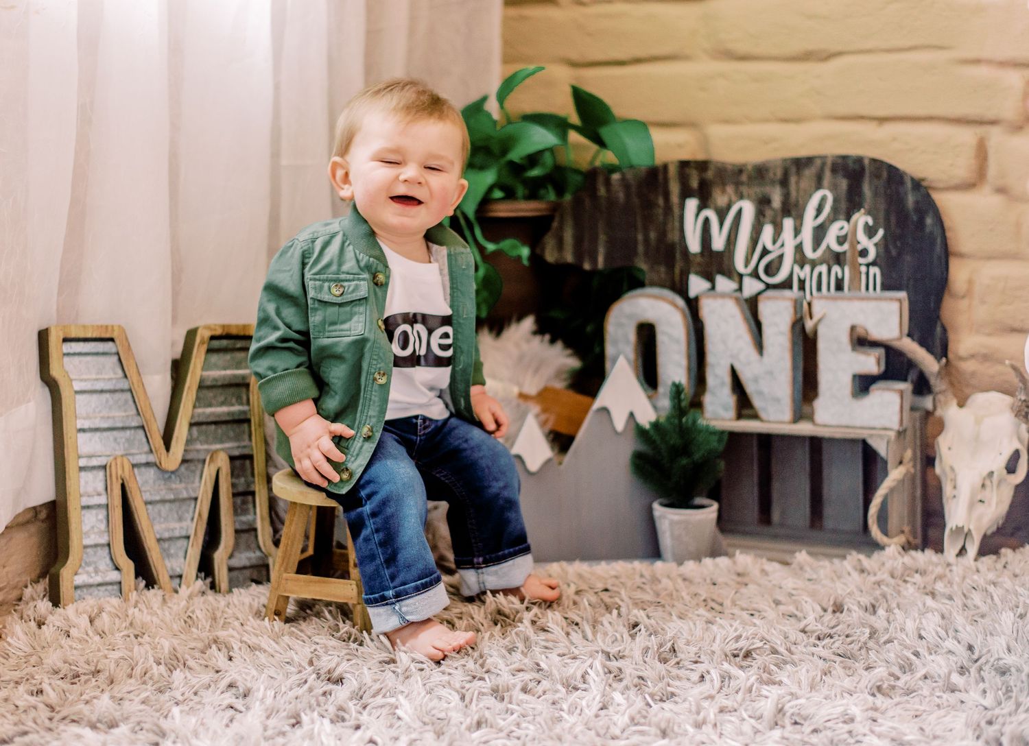 First birthday photo setup with rustic decorations including the word ONE, letter M, and a plush gray rug.
