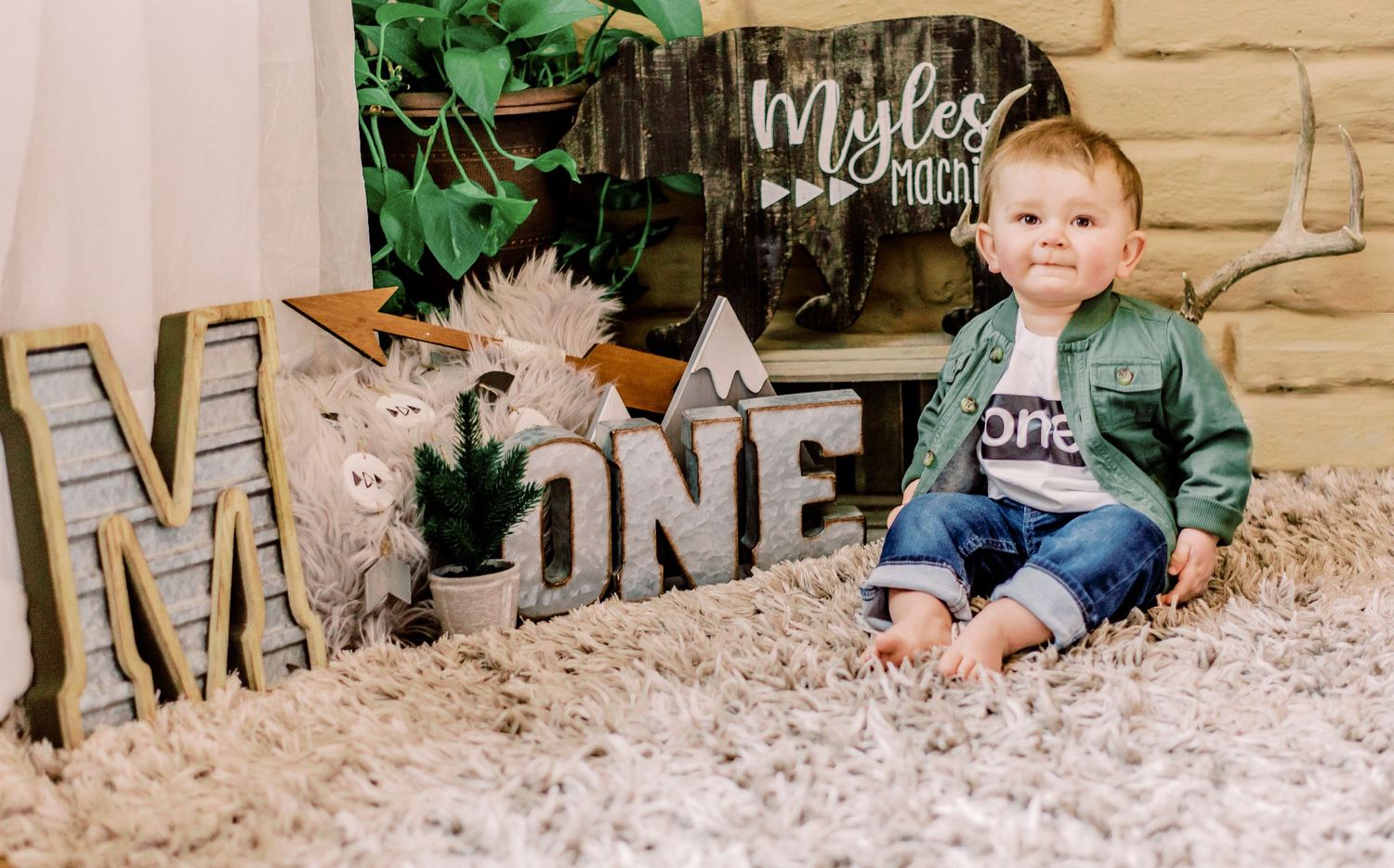 A rustic first birthday scene with silver "ONE" letters, soft rug, and floral decorations against a wooden backdrop.