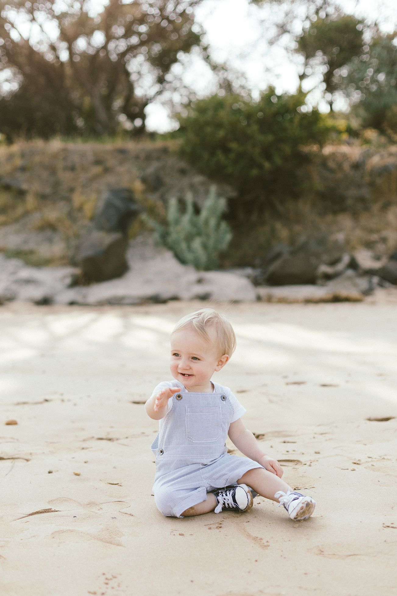A baby in white romper and sneakers sits on a sandy beach near desert plants.
