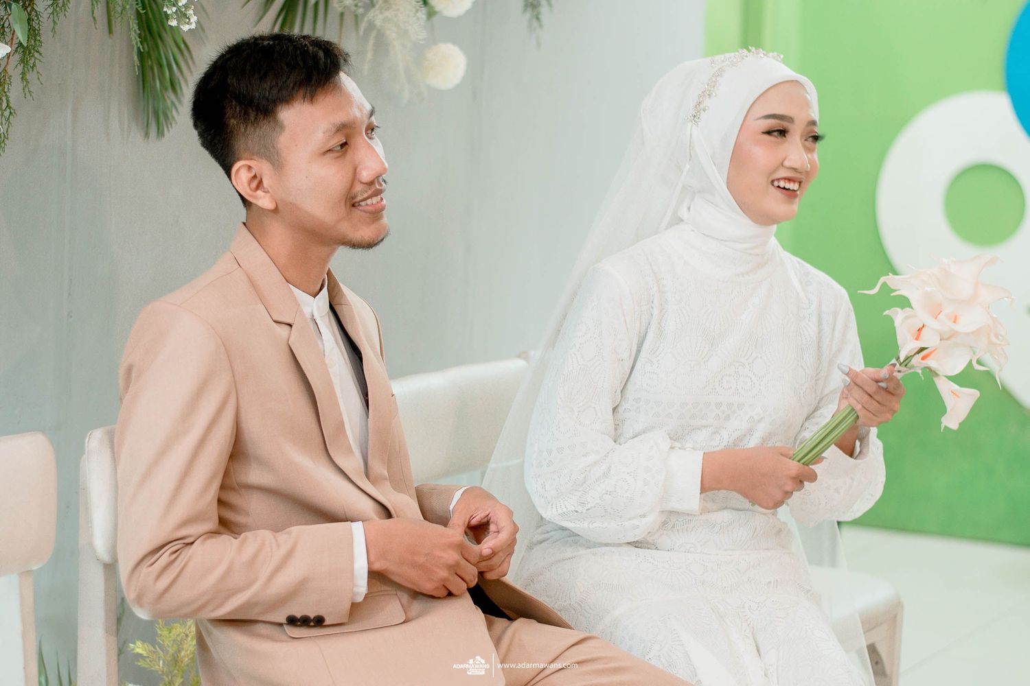 A couple in coordinated beige and white outfits sits together during their wedding celebration.