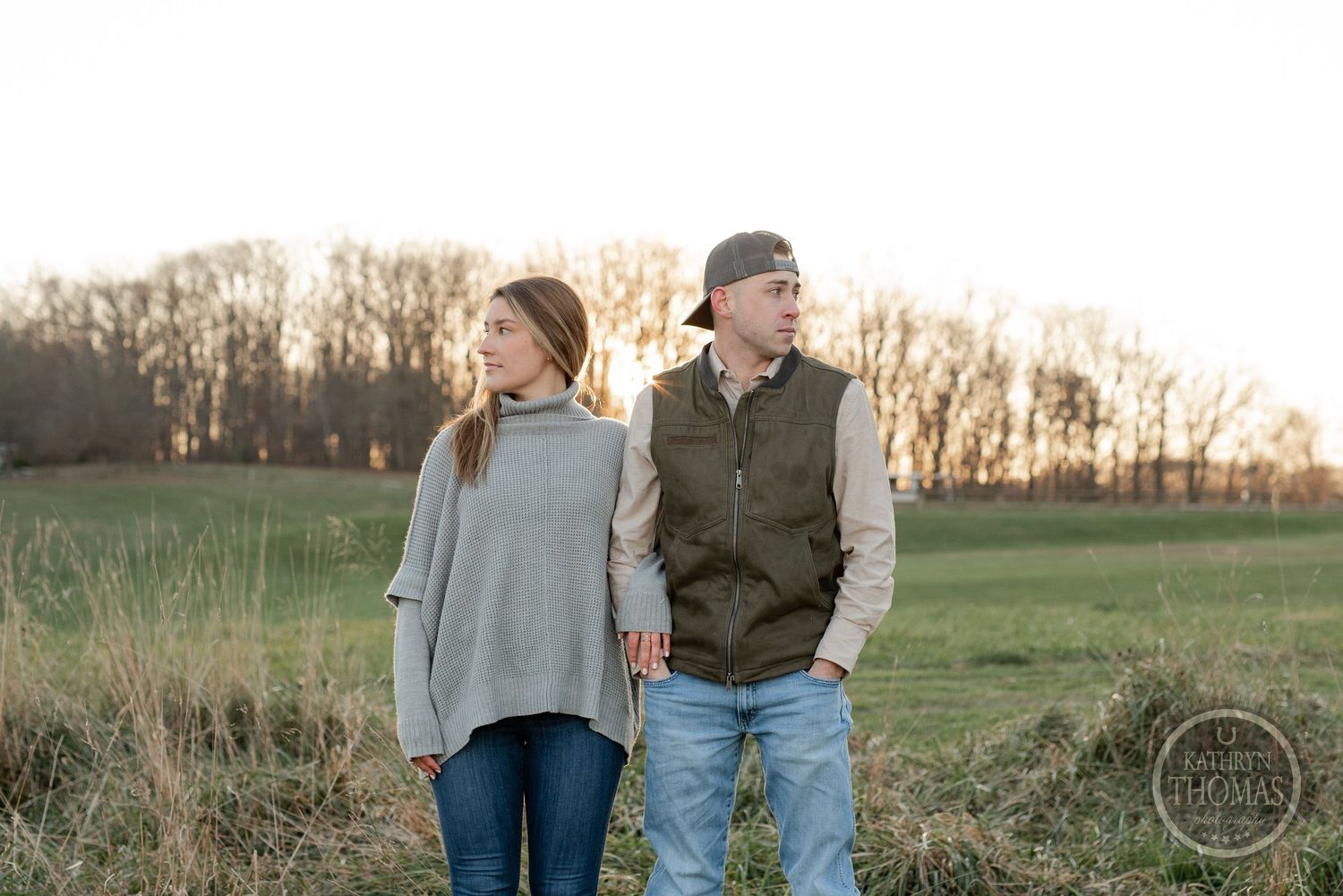 A couple in casual attire stands together in a serene rural field at sunset.
