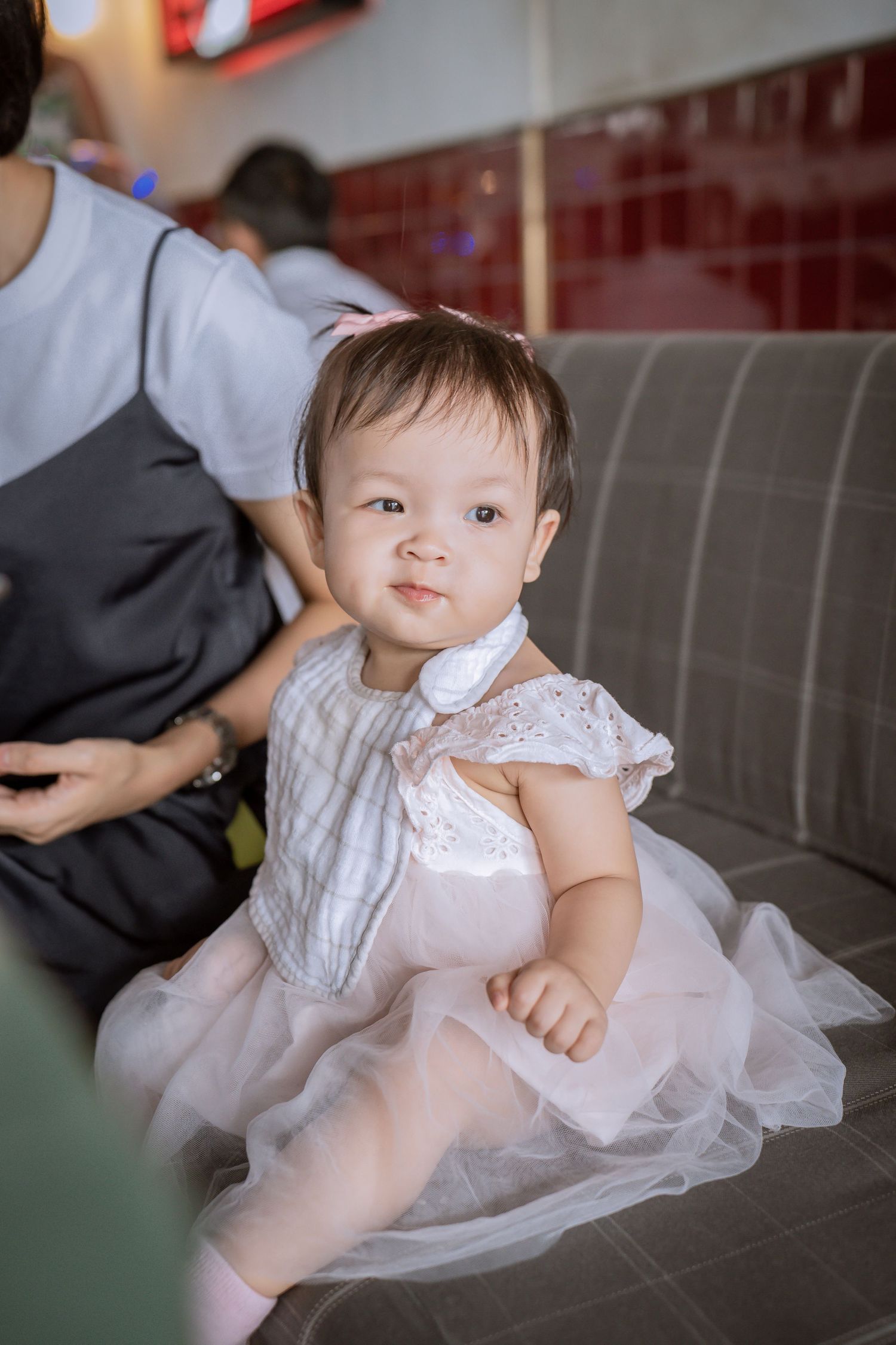A baby wearing a white tulle dress sits on a grey couch.