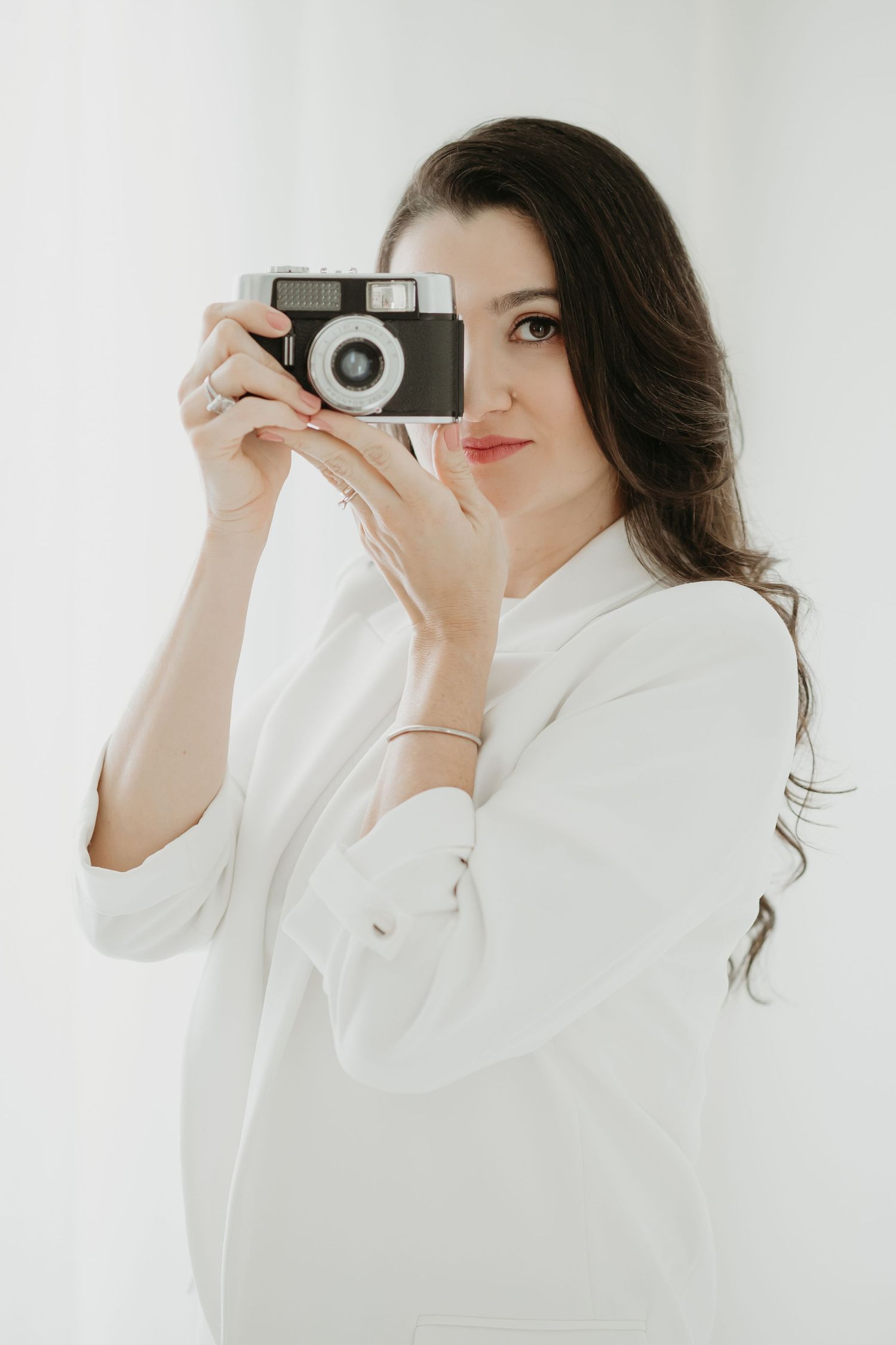 Photographer in white blazer holding vintage camera against bright studio backdrop.