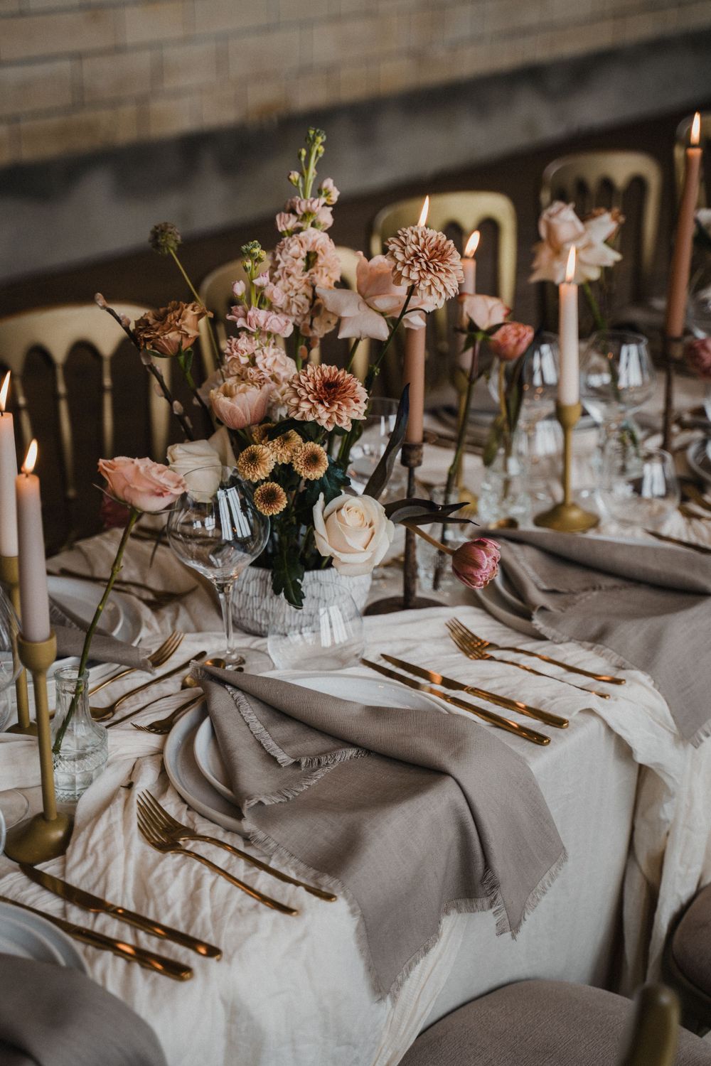 Elegant wedding table setting with muted pink and white floral centerpiece, gold cutlery, candles, and neutral linens on rustic table.