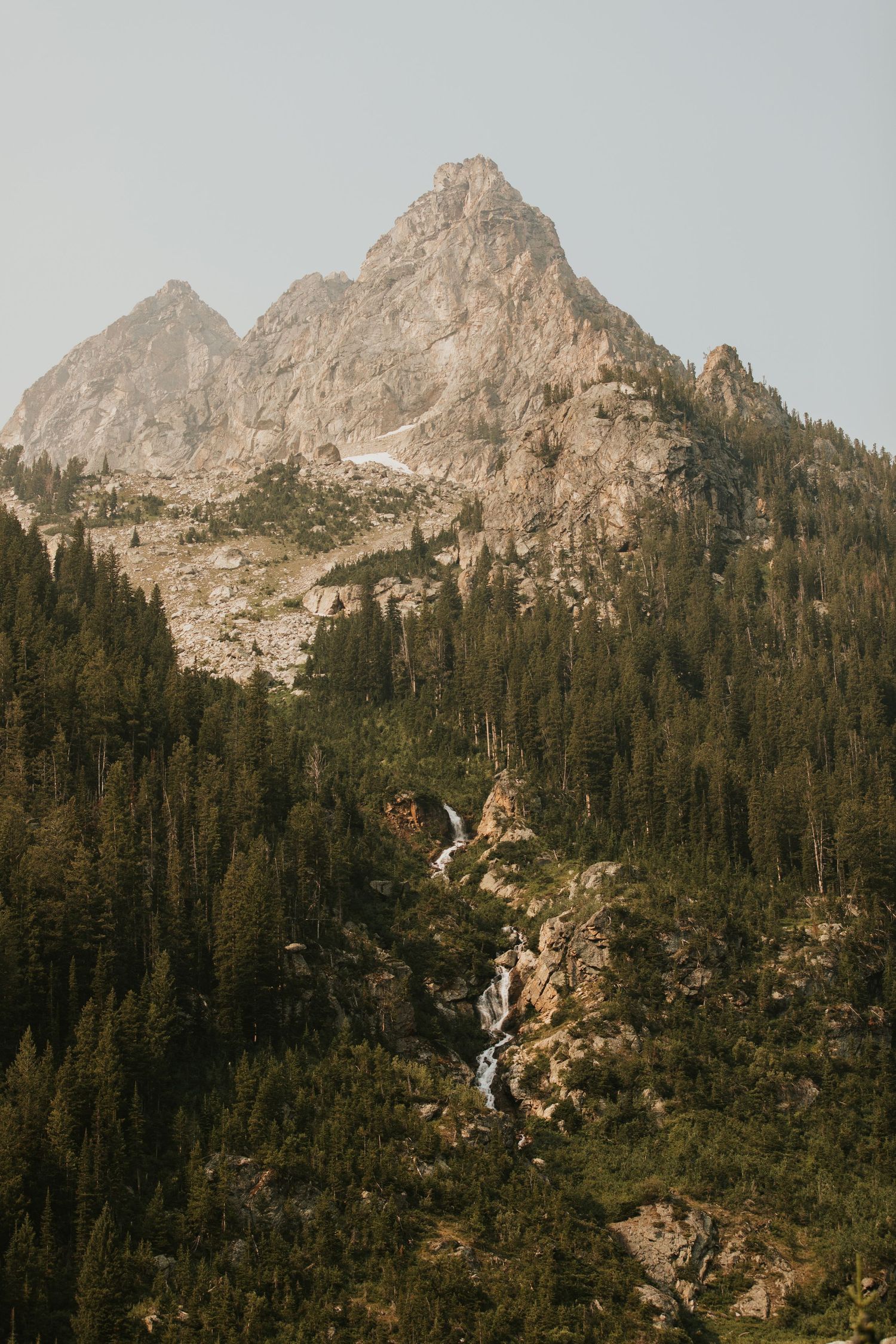Majestic mountain peaks rise above dense forest with cascading waterfall in wilderness landscape.