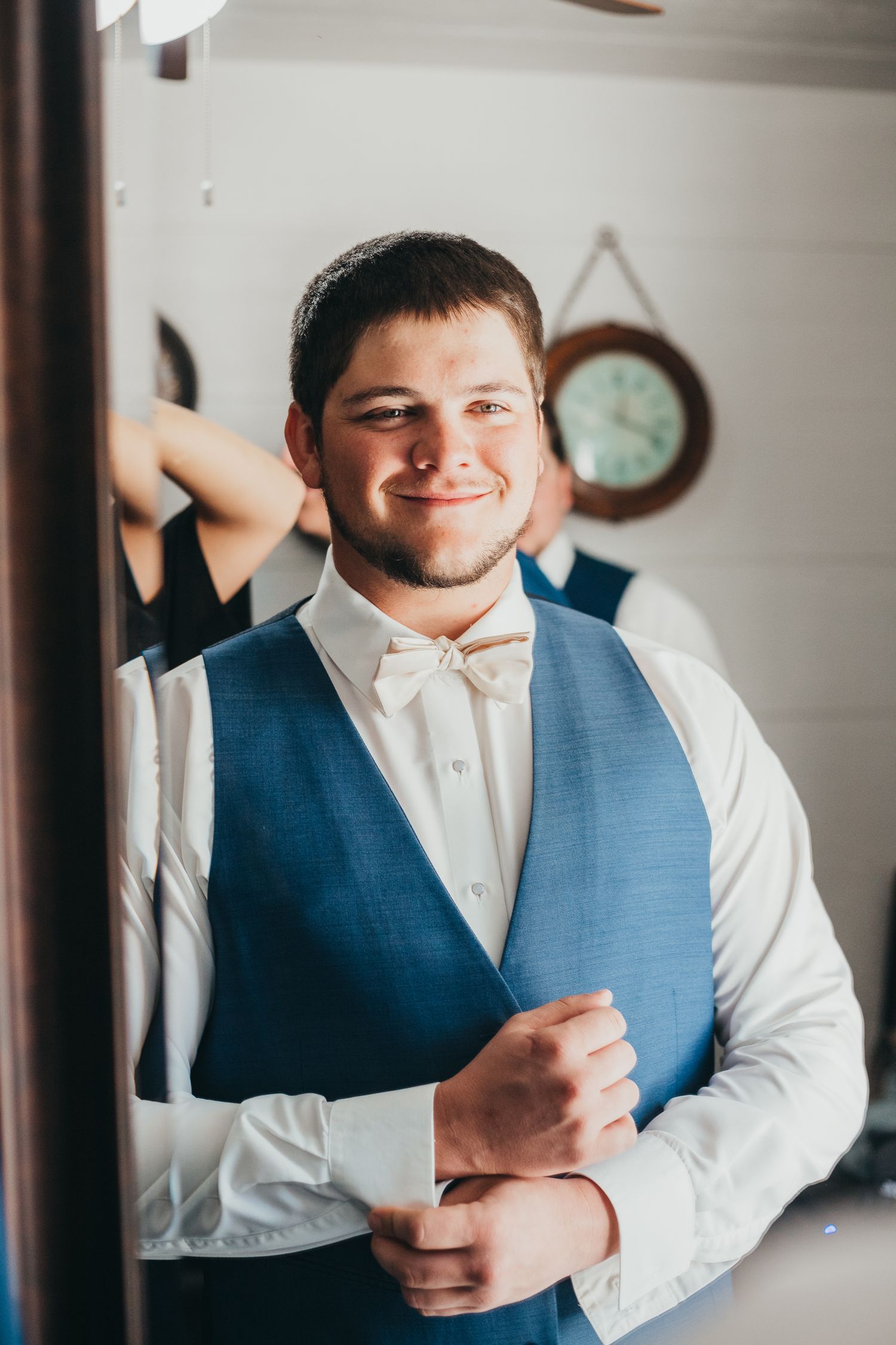 Someone in formal attire wearing a white dress shirt, light blue vest and bow tie smiles while adjusting their cuff.