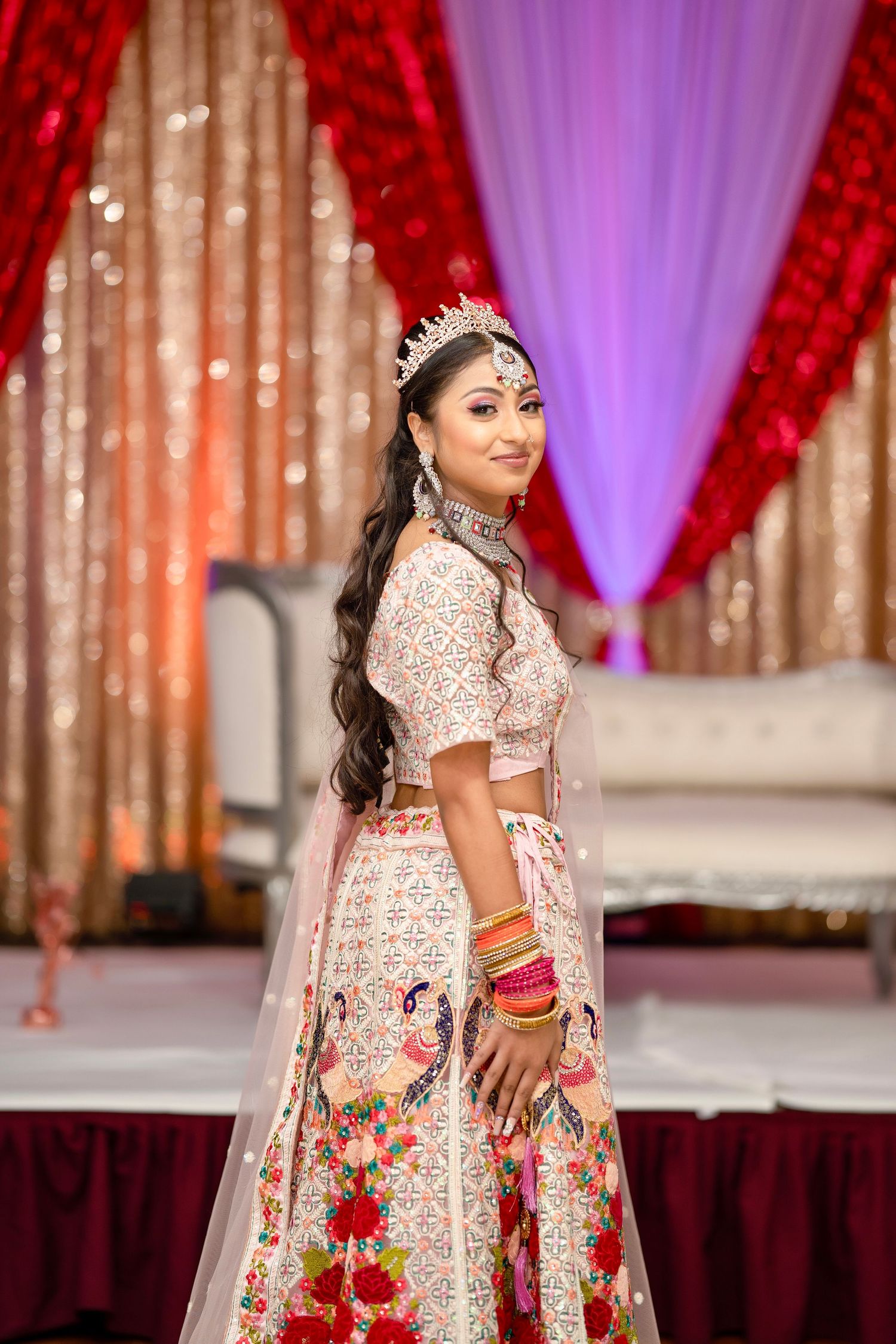 Traditional Indian bride in pink floral lehenga poses against sparkly backdrop with red drapes.