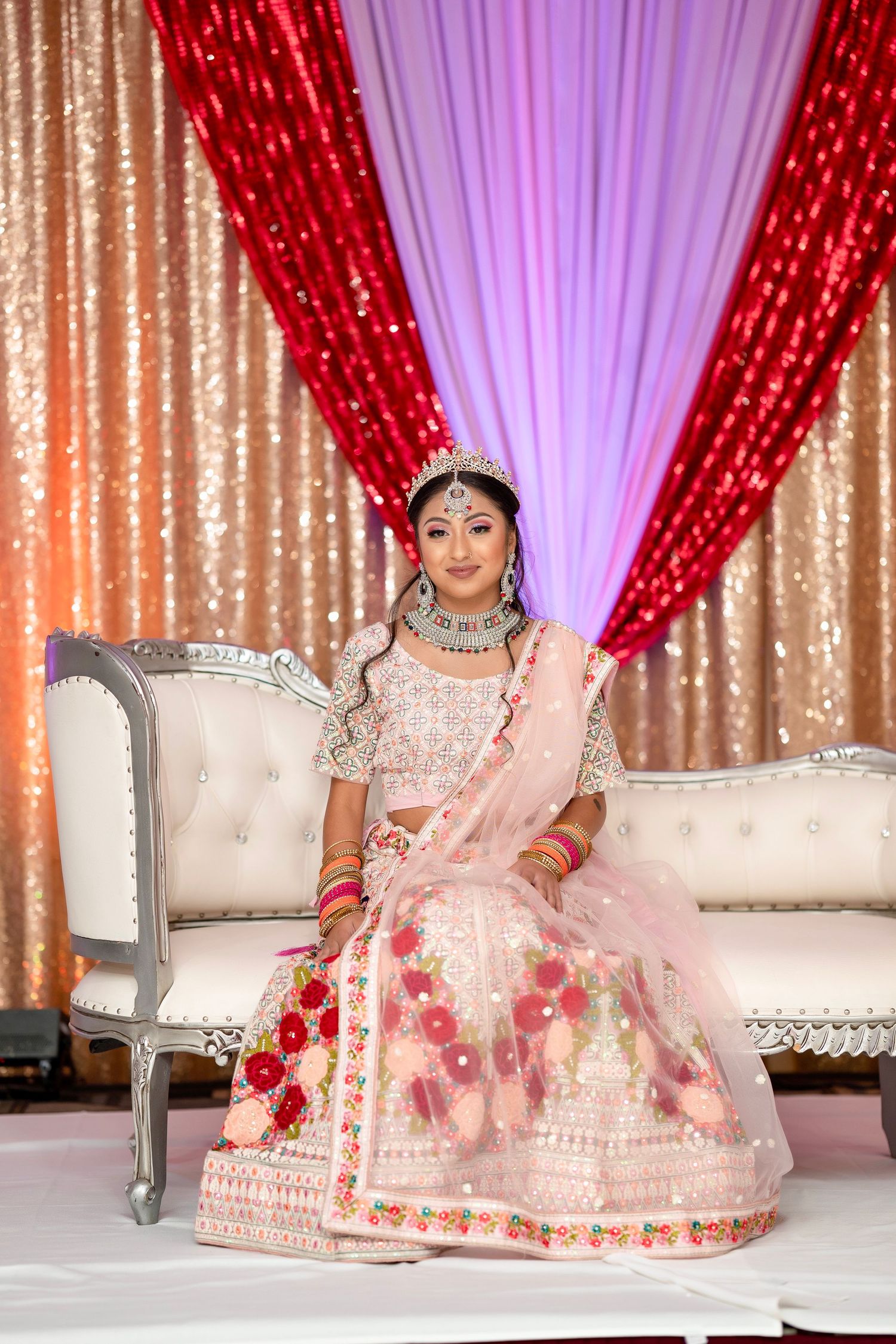 Elegant bridal portrait on ornate sofa against red and gold sequined backdrop with purple draping.
