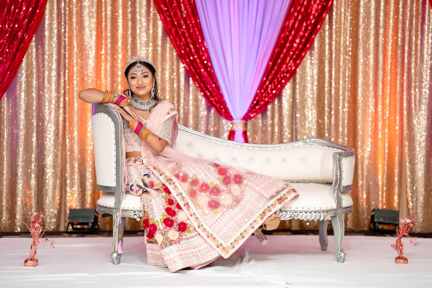 Bride in embroidered lehenga reclines on white chaise against glittering backdrop.