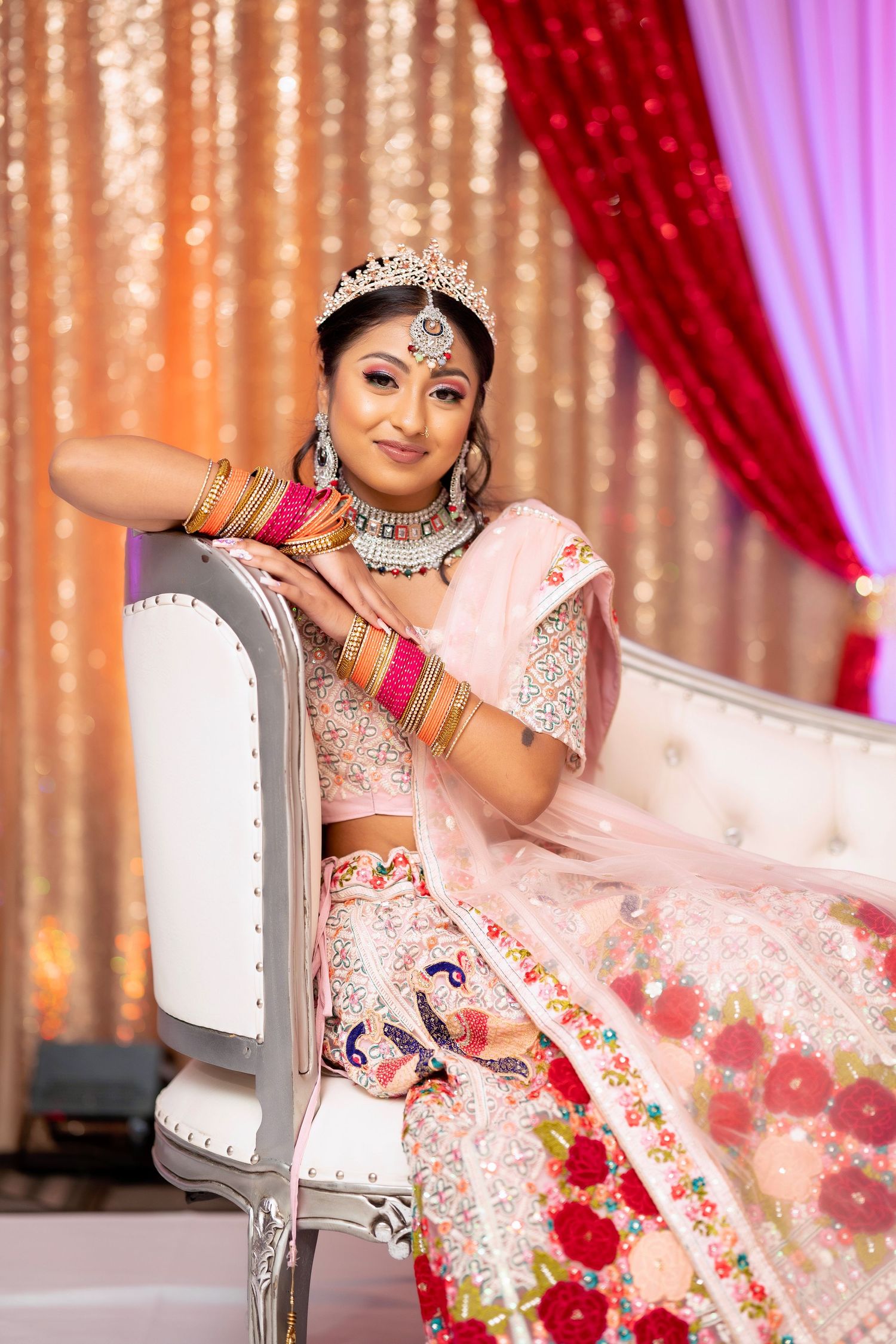 Traditional bridal pose in ornate pink lehenga while seated on white chair with decorative backdrop.