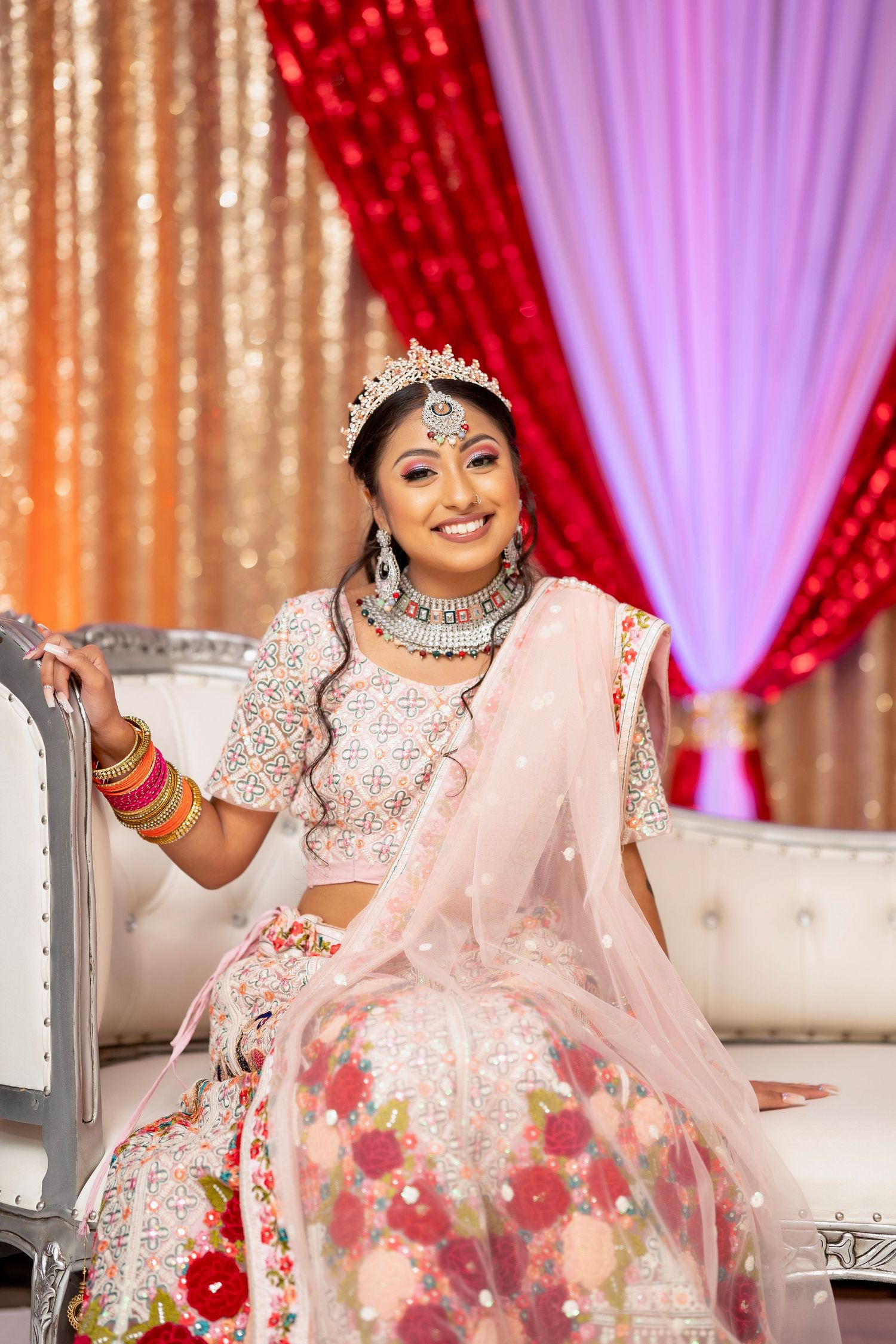 Smiling bride in pink floral lehenga and crown poses on elegant white sofa.