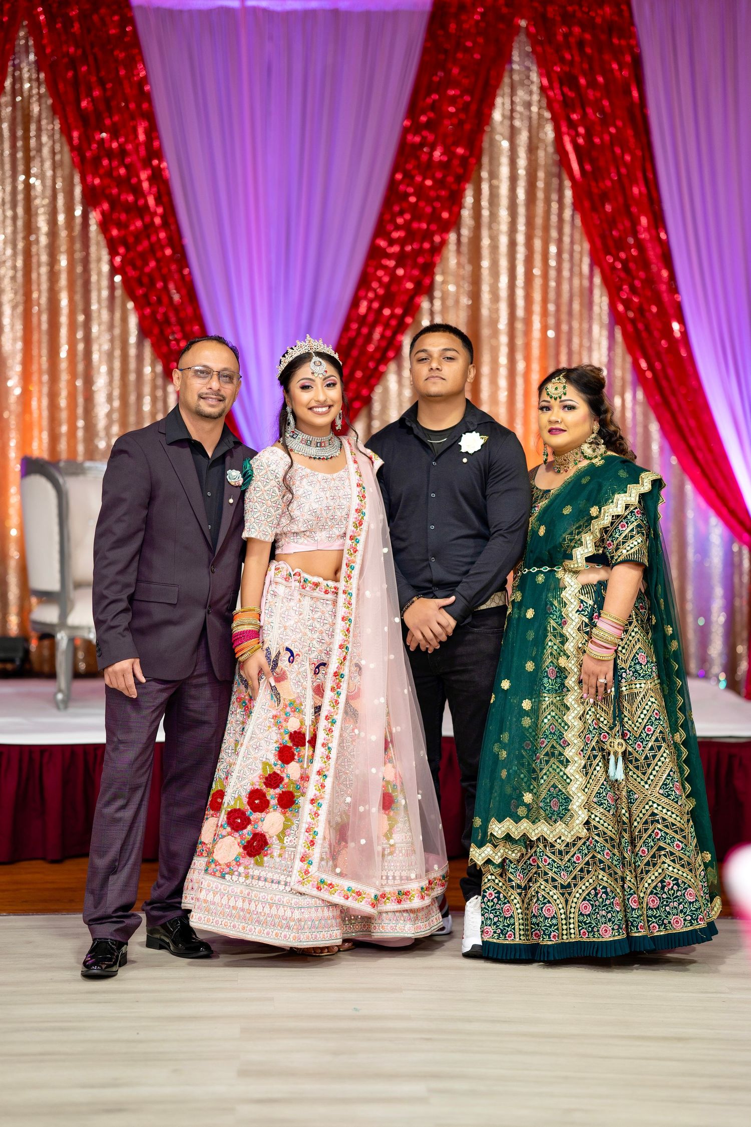 Family group photo at celebration event with traditional Indian formal wear against red and gold backdrop.