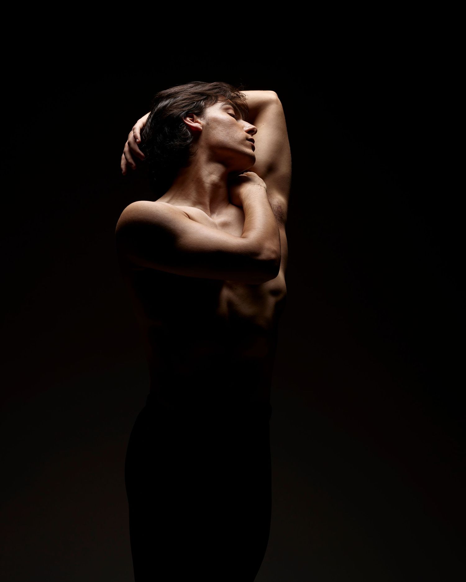 Dramatic low-key portrait showing silhouette of figure in dark clothing against black background.