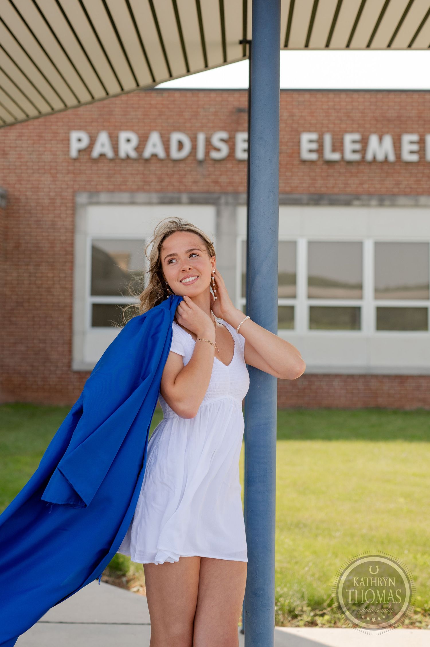 Graduate in blue graduation gown and white dress poses near Paradise Elementary entrance.