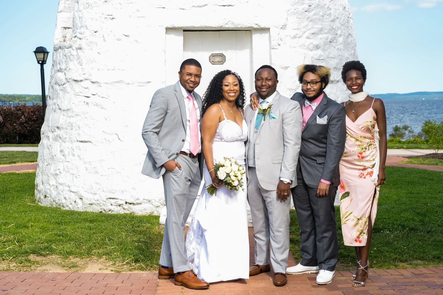 A wedding party poses in front of a white lighthouse on a sunny day by the ocean.