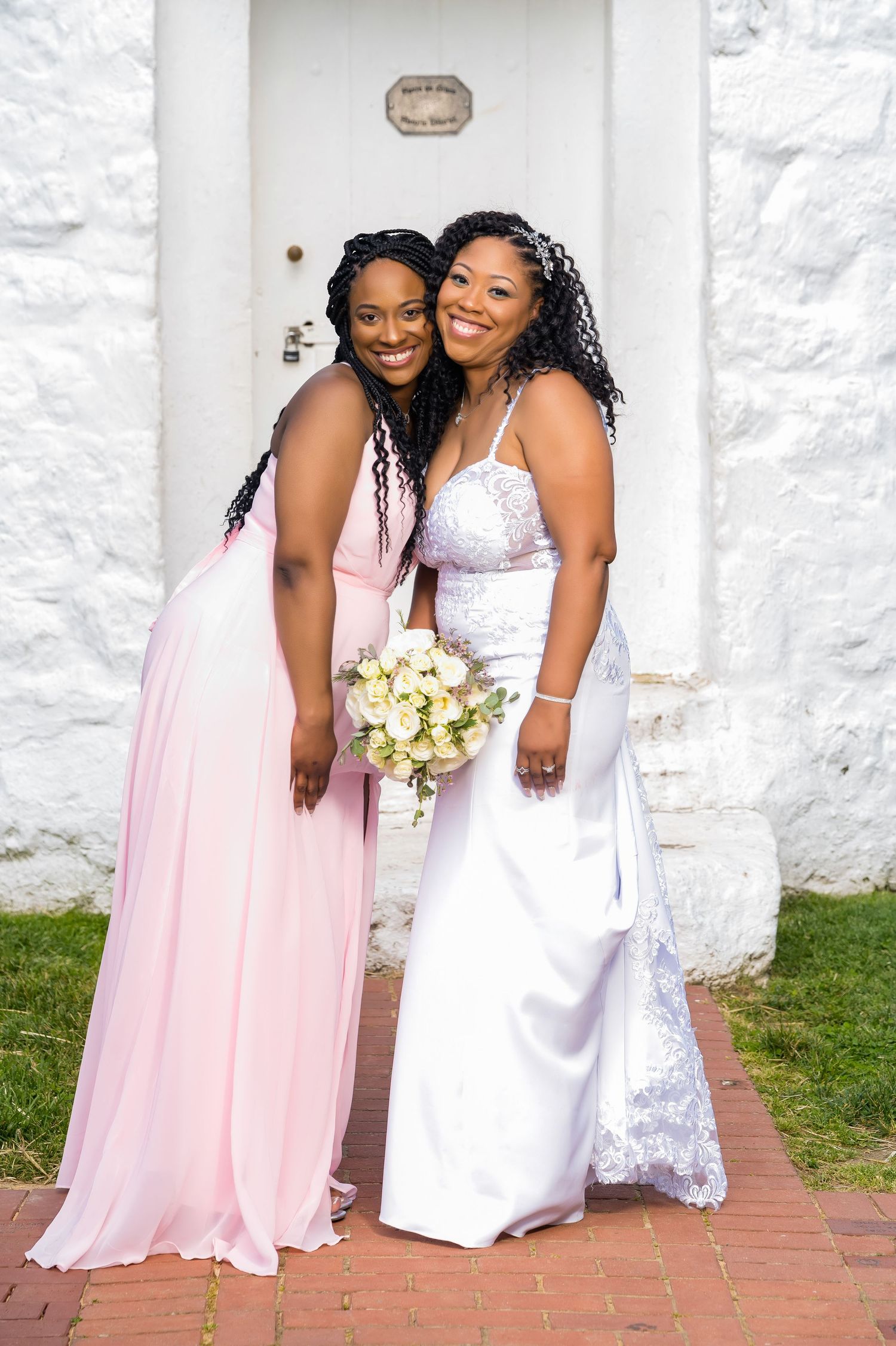 A bride in a white lace gown poses with bridesmaid in pink flowing dress holding a bouquet at a white church doorway.