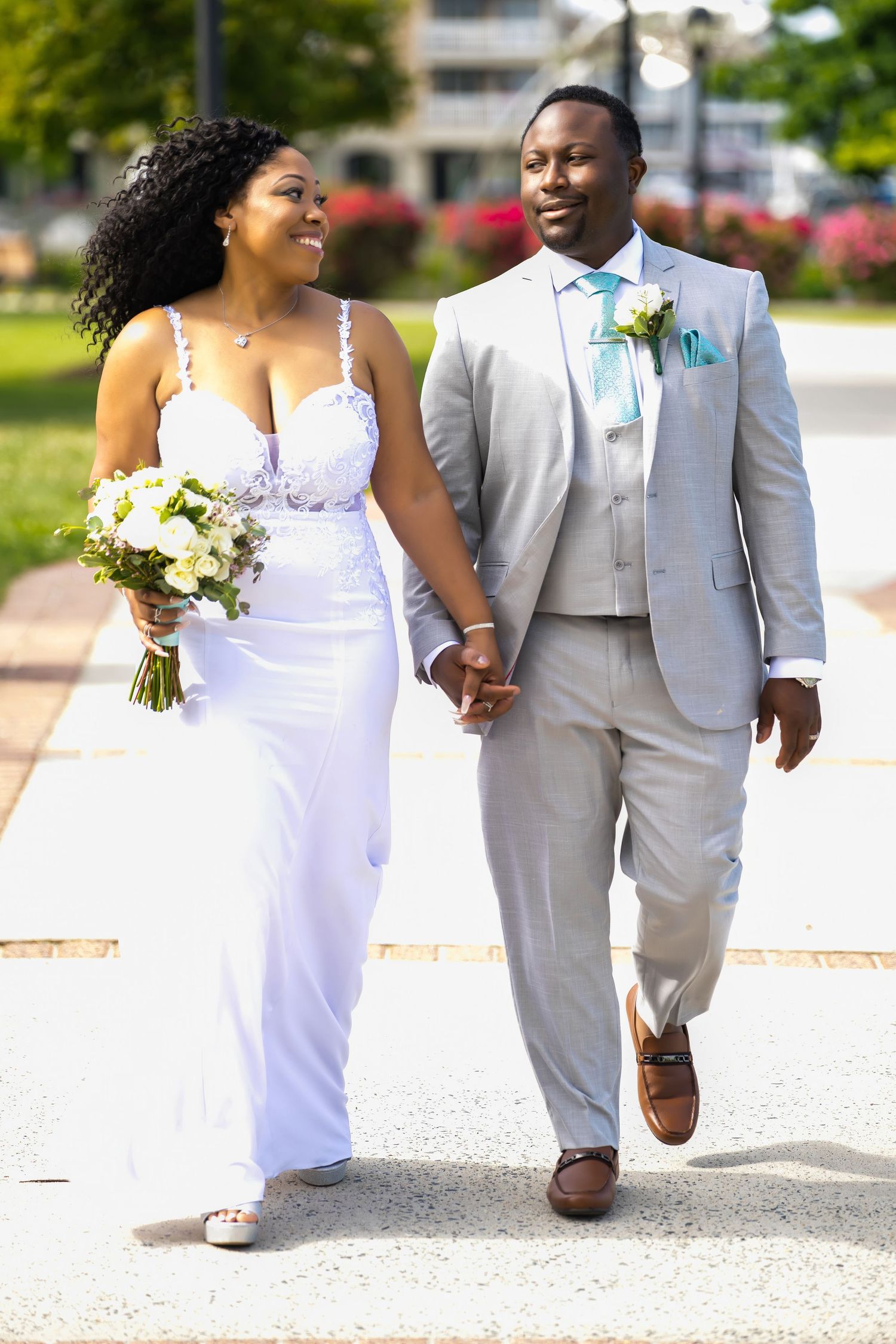 A joyful couple walks hand in hand on their wedding day, wearing a white gown and light grey suit in a garden setting.
