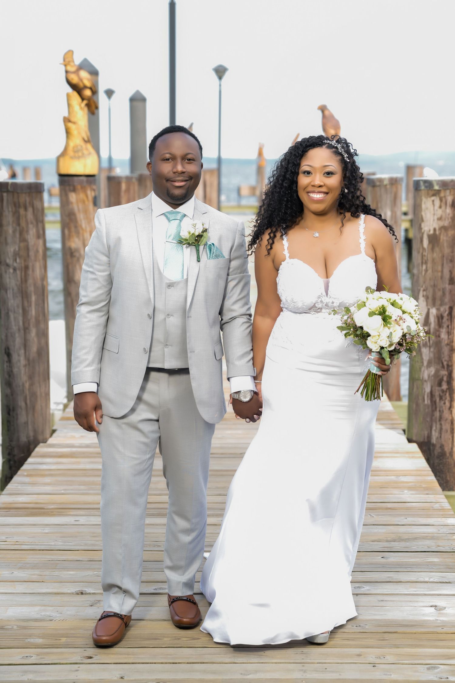 vA joyful couple walks hand in hand on their wedding day, wearing a white gown and light grey suit in a garden setting.