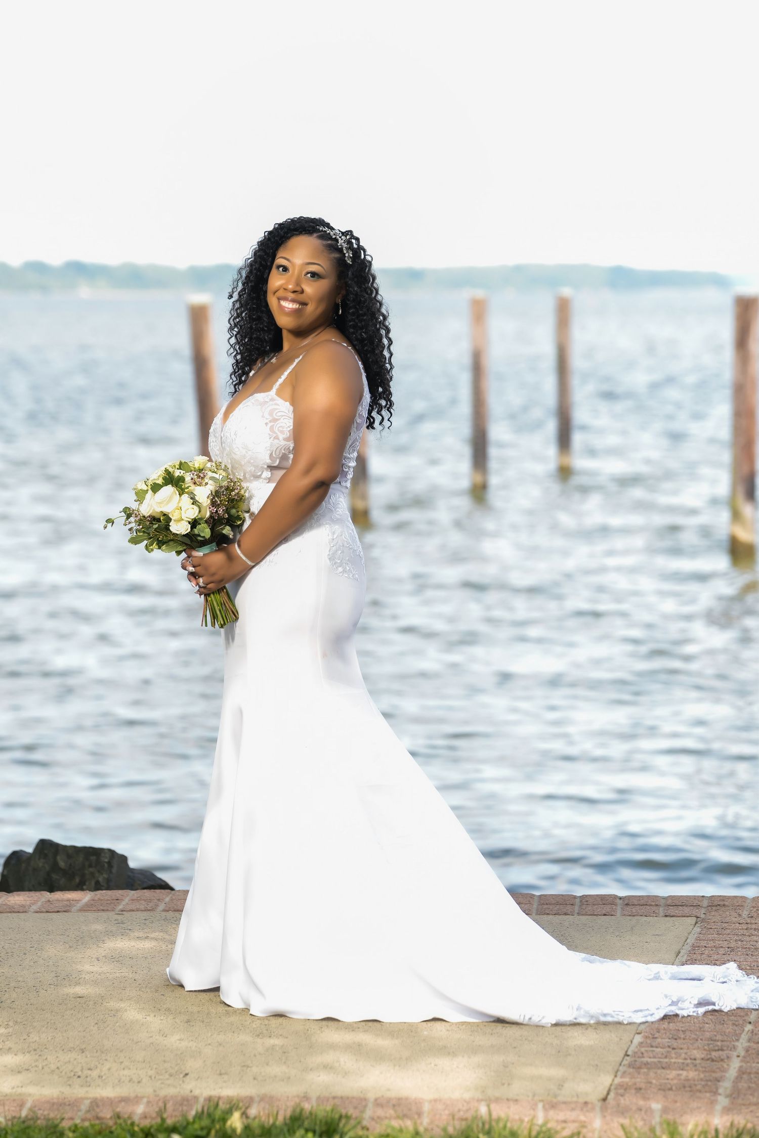 A bride in a stunning white mermaid wedding dress holds a bouquet while posing by the waterfront with wooden posts behind her.
