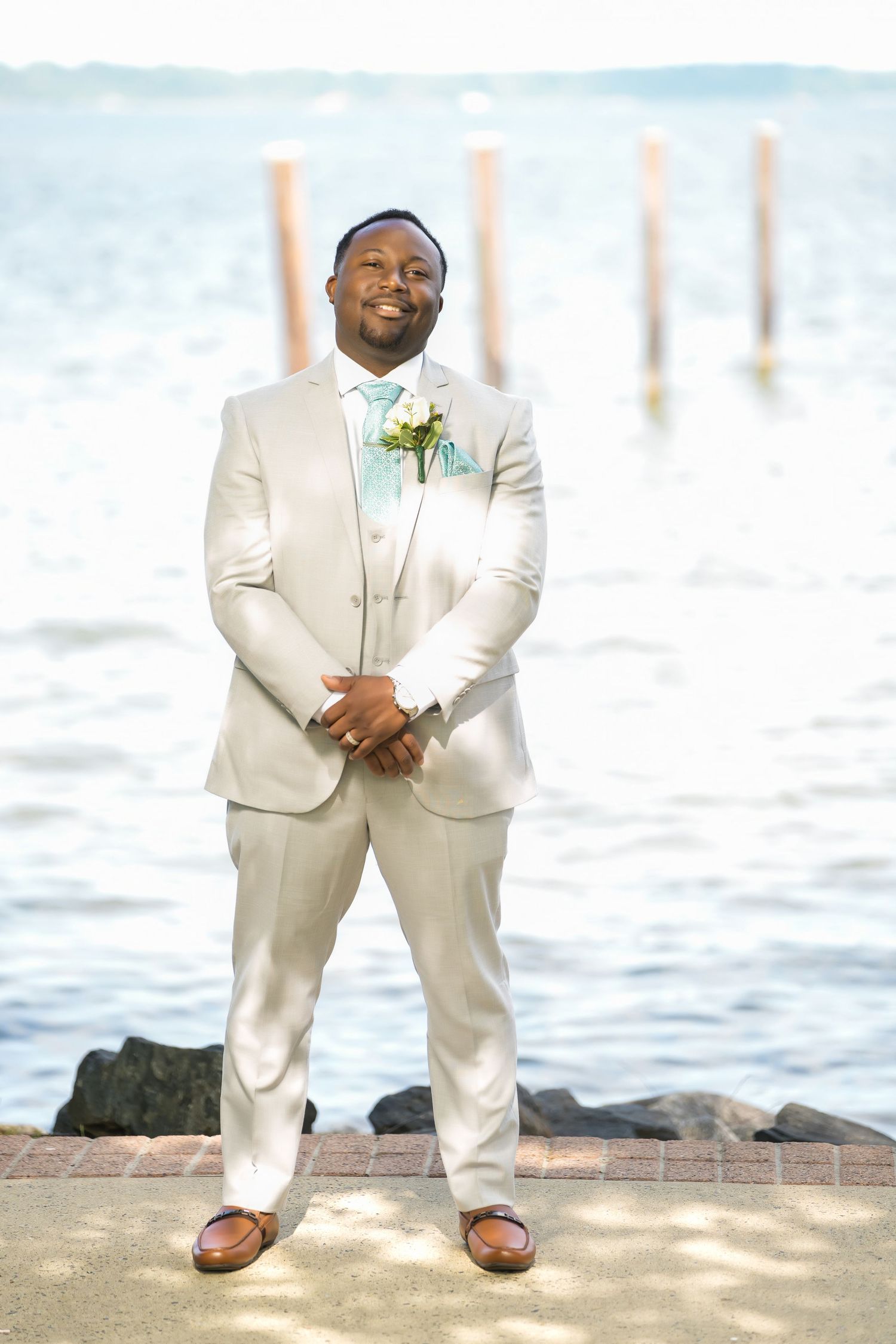 A person in a cream-colored wedding suit with a mint green bowtie stands on a pier by the ocean.