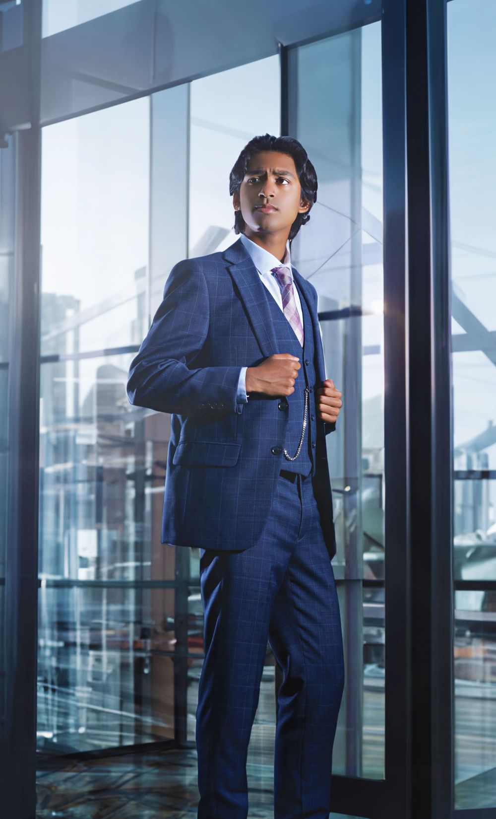 Professional in navy blue business suit stands confidently by office window with city skyline backdrop.