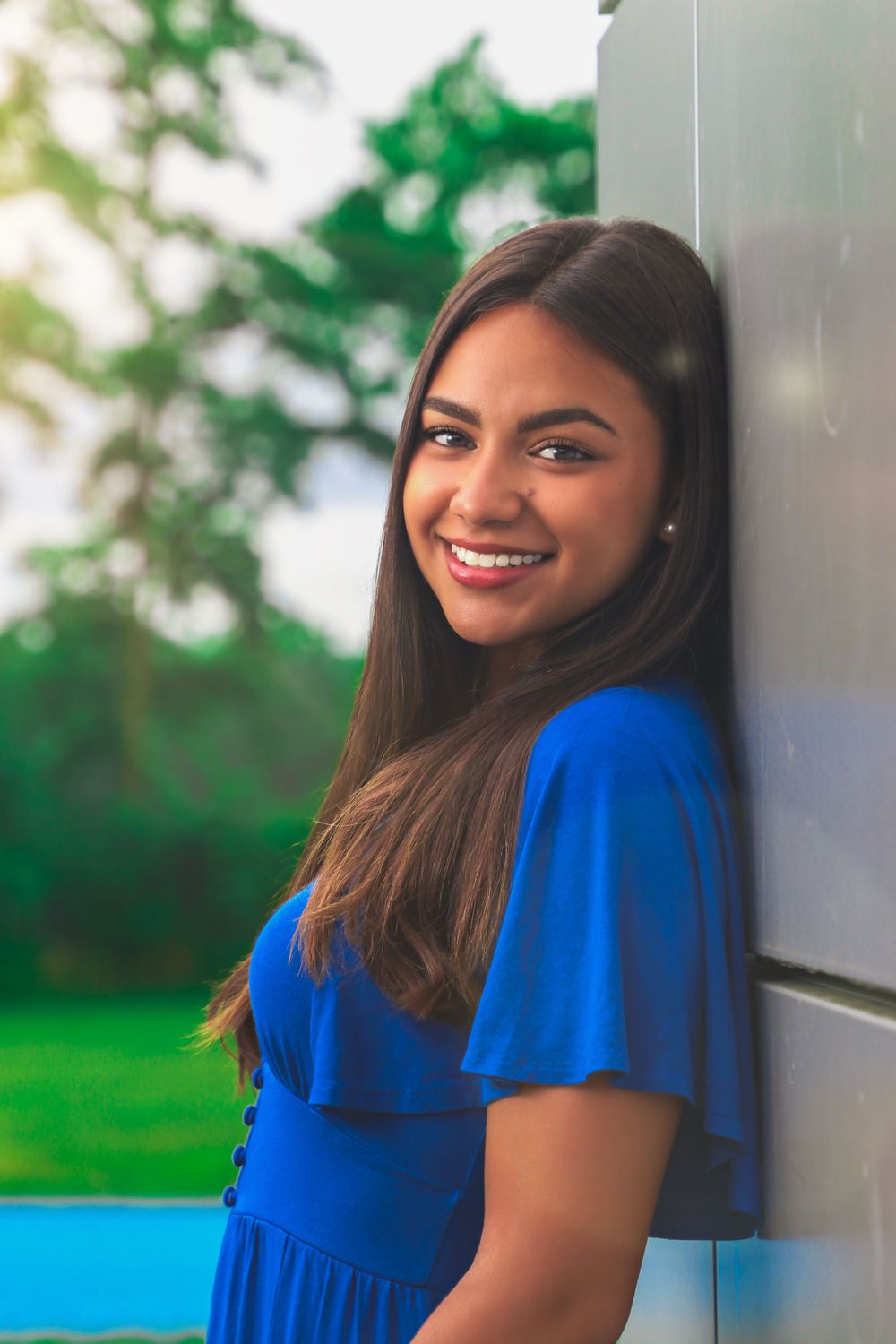 Portrait of a person in vibrant blue top smiling against outdoor setting with greenery.