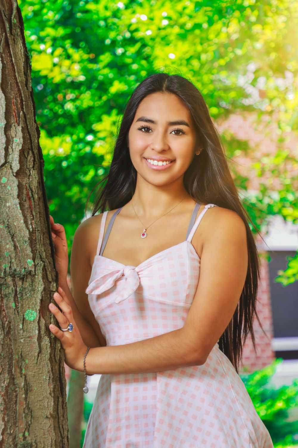 Portrait in pink gingham dress leaning against tree trunk with green foliage background.