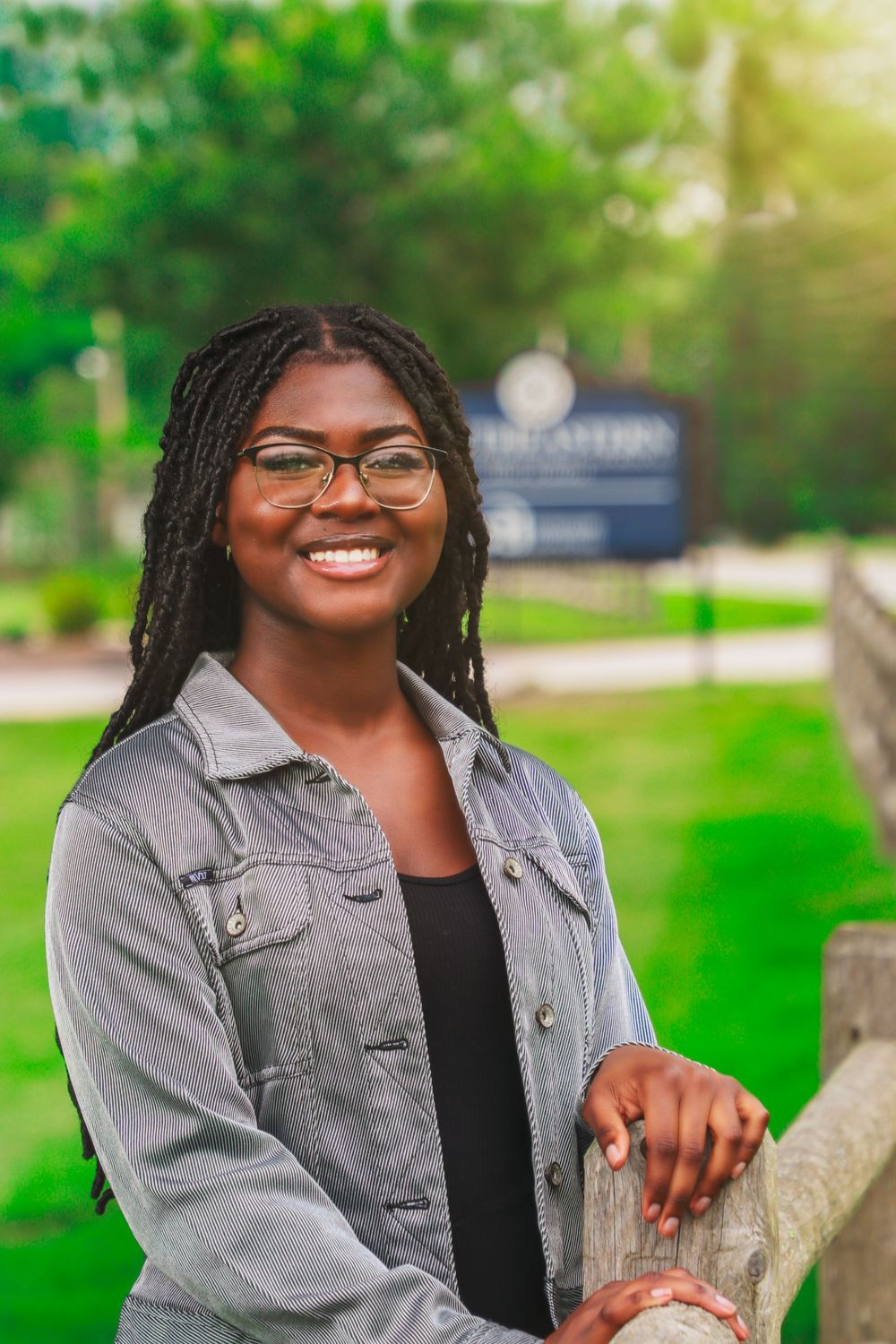 Portrait wearing denim jacket and glasses while leaning on wooden fence in park setting.