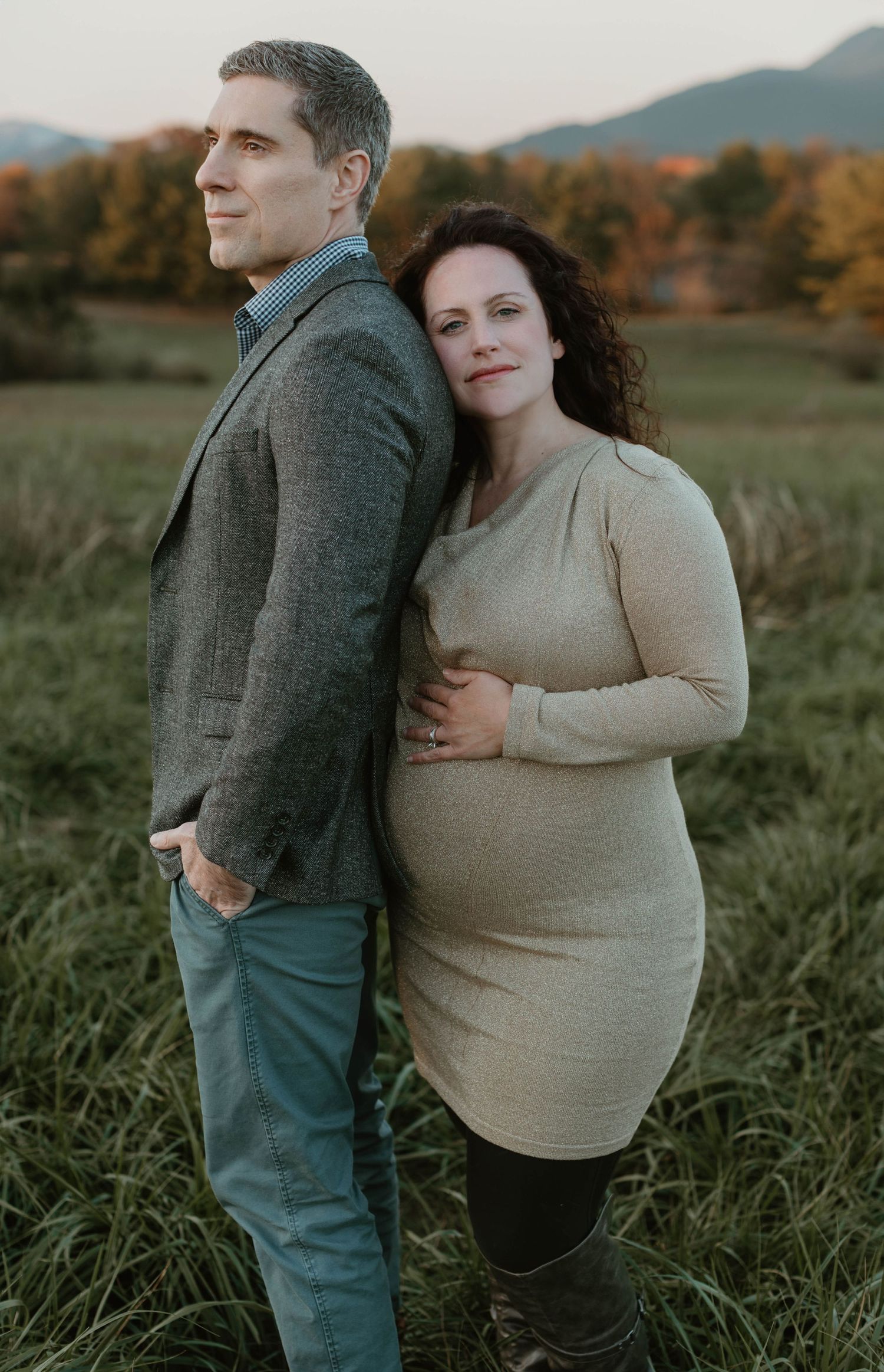 Couple embraces in a field during sunset with mountains in background and autumn foliage.