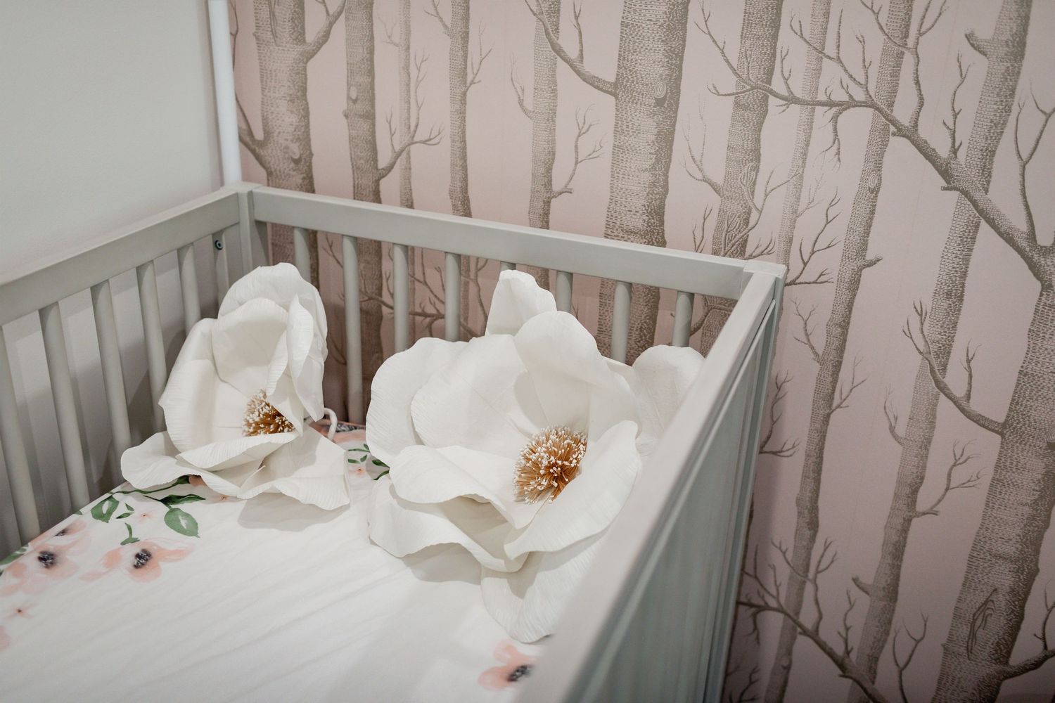 Large white floral decorations adorn a gray crib with birch tree wallpaper backdrop.