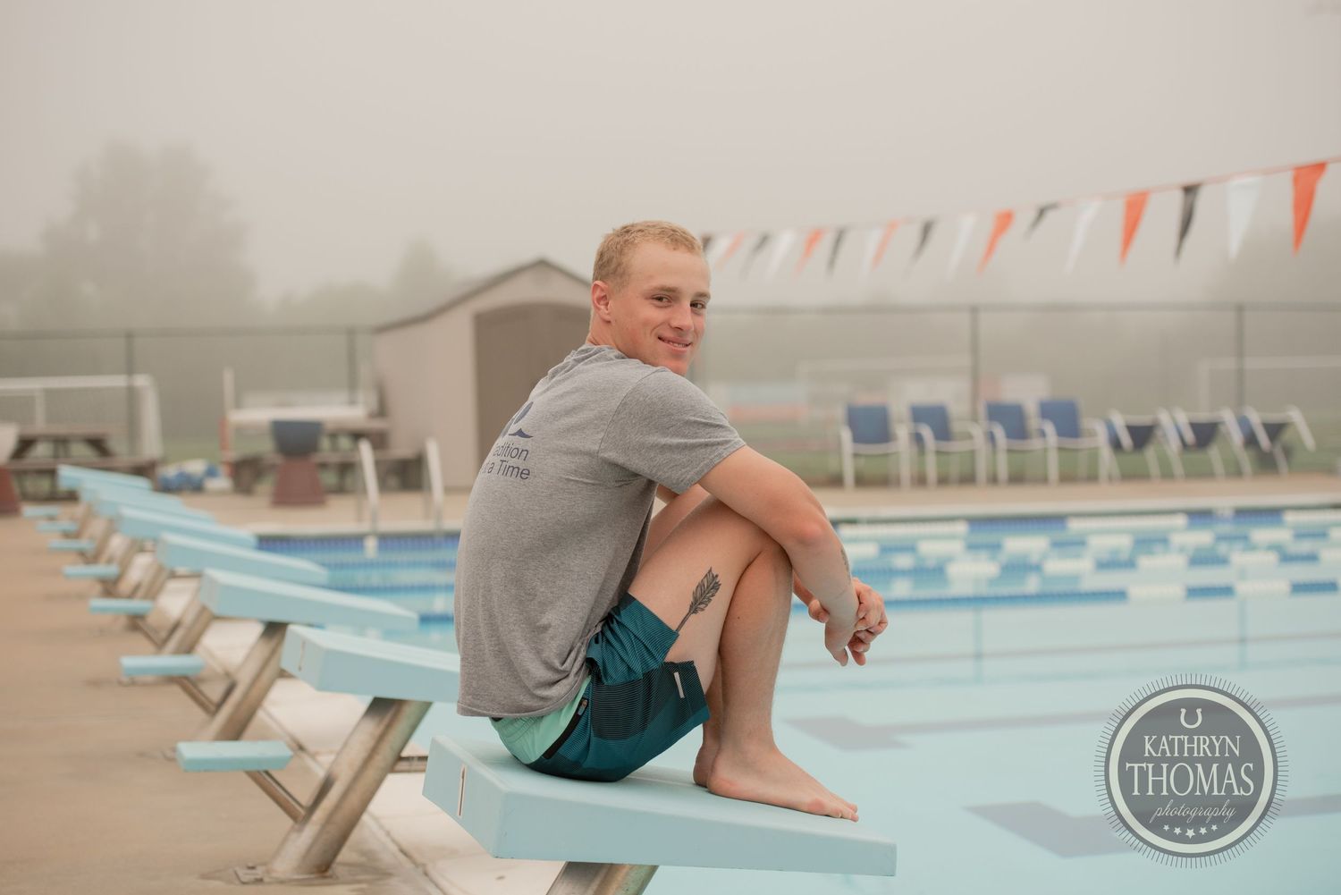 A swimmer in gray shirt sits on diving board above pool on foggy morning.