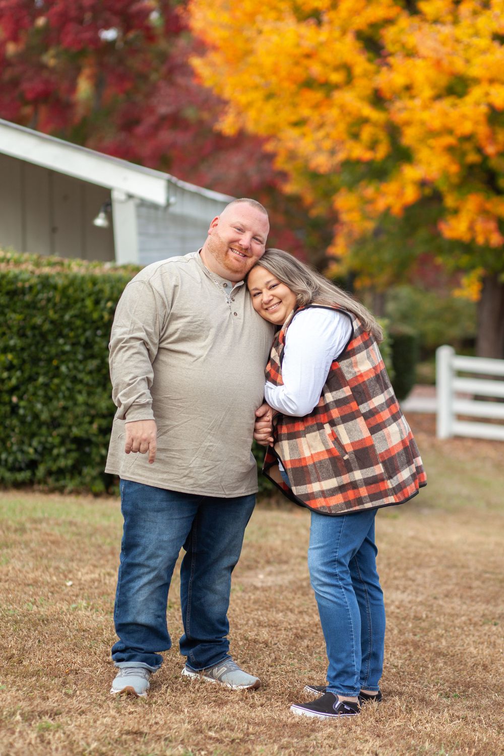 Couple embracing outdoors in autumn with vibrant fall foliage and white fence in background.
