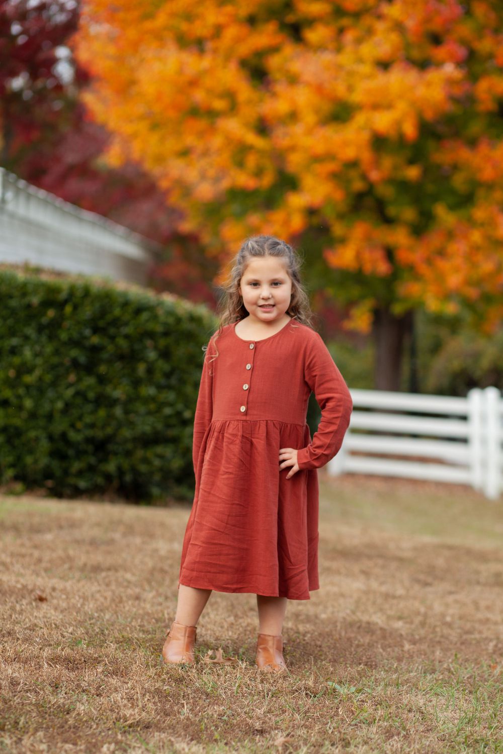 Child wearing a rustic red dress stands in front of autumn foliage and white fence on brown grass.
