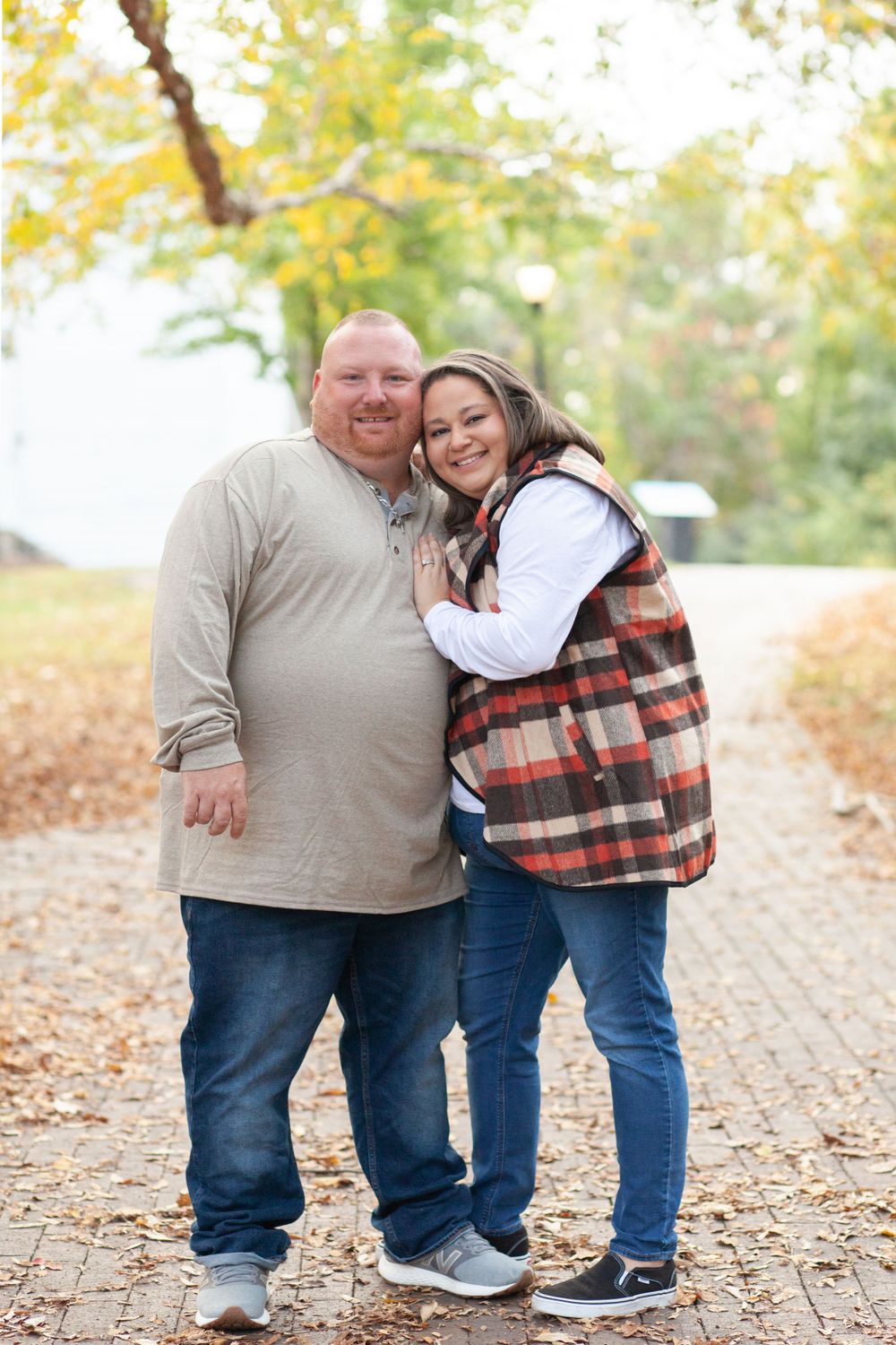 A couple smiles together on an autumn path with fallen leaves and yellow foliage in the background.