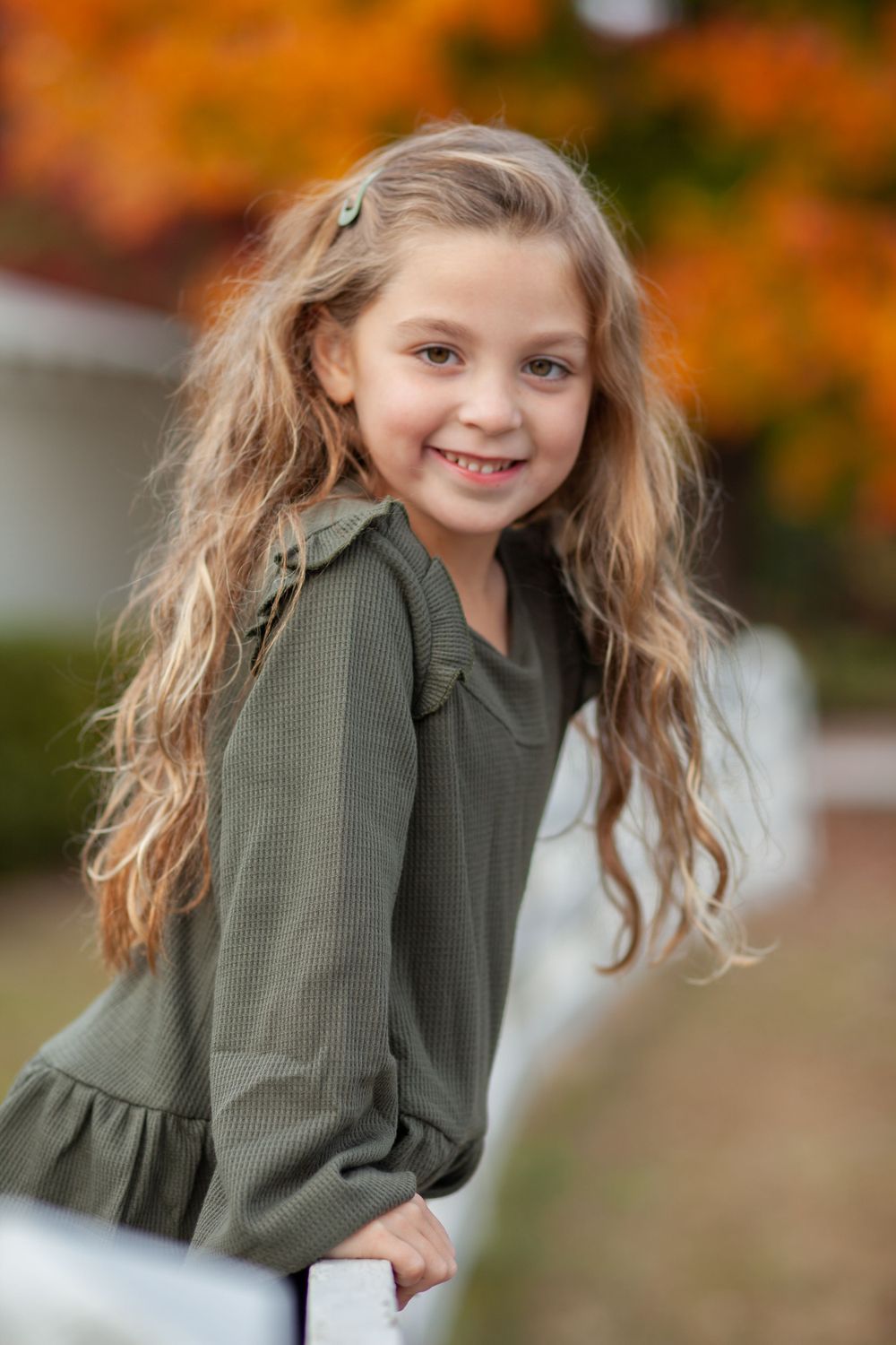 A young person wearing a sage green ruffled top poses outdoors against vibrant autumn foliage in the background.