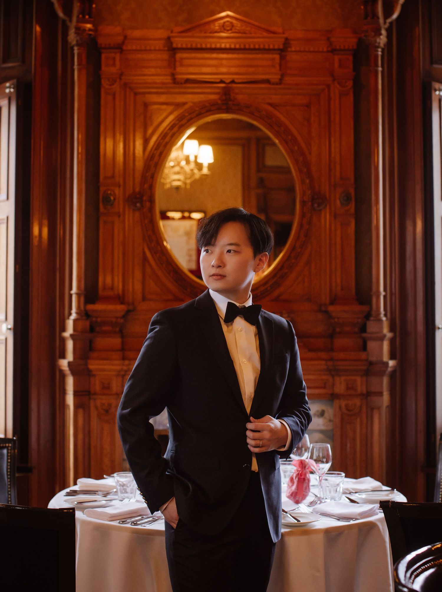 Groom in classic tuxedo posing by elegant round mirror and dining setup.