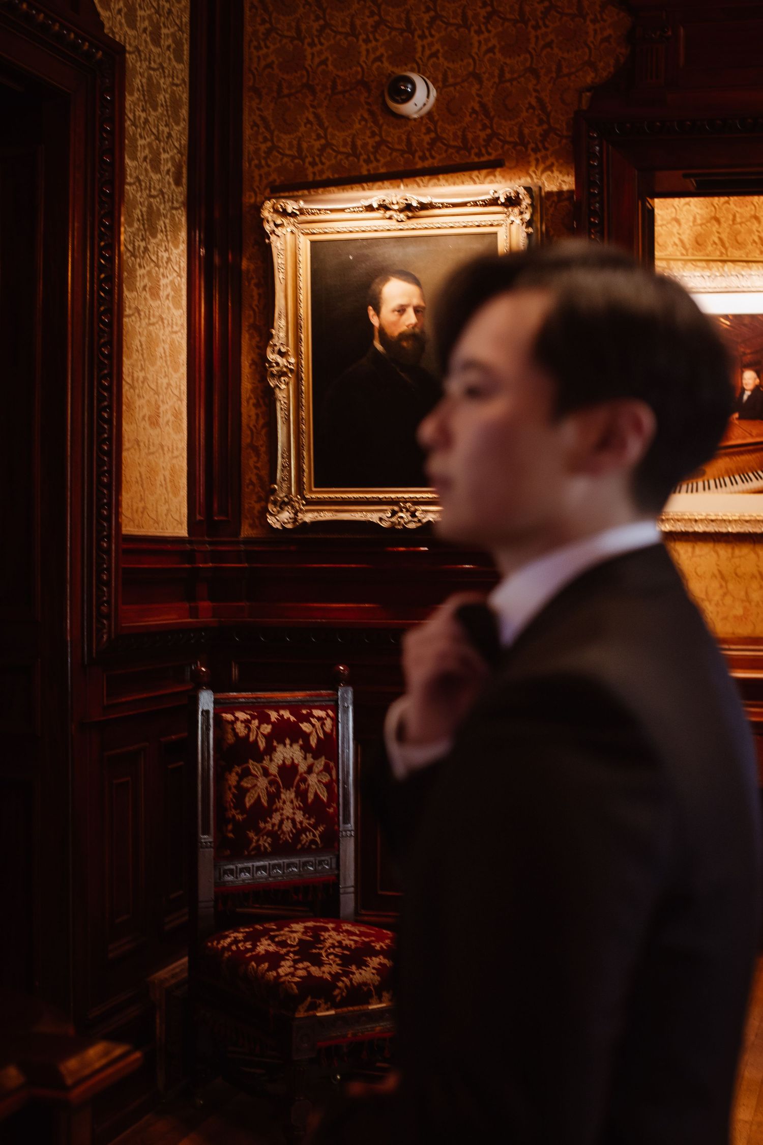 Groom adjusting tie with vintage portrait in ornate frame behind.