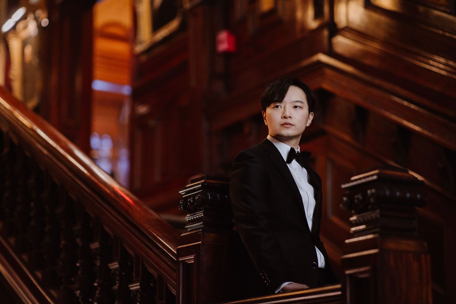 Stylish groom stands on vintage wooden stairs in classic black tuxedo.