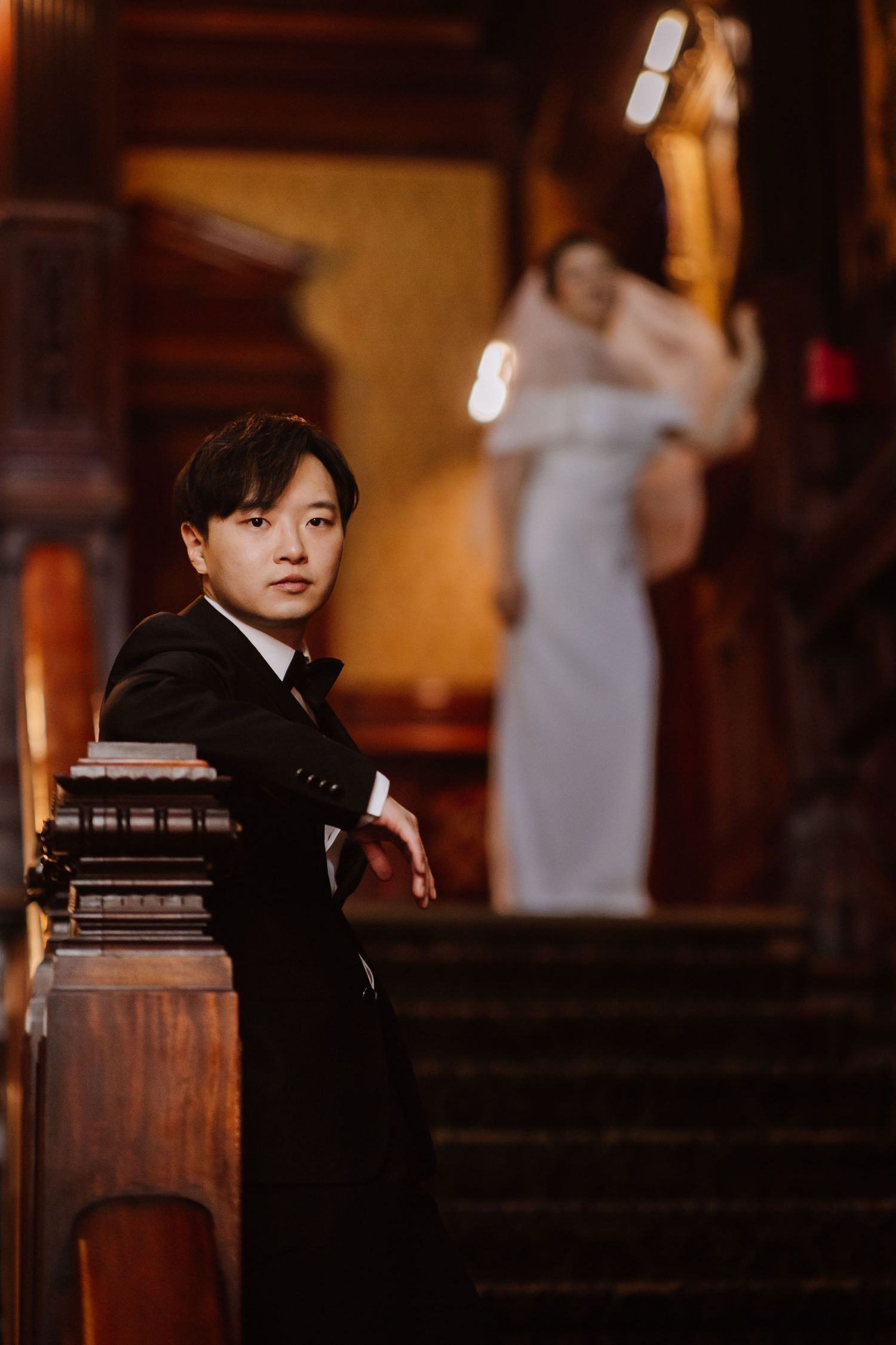 Elegant groom in black tuxedo poses against ornate wooden staircase in warm lighting.
