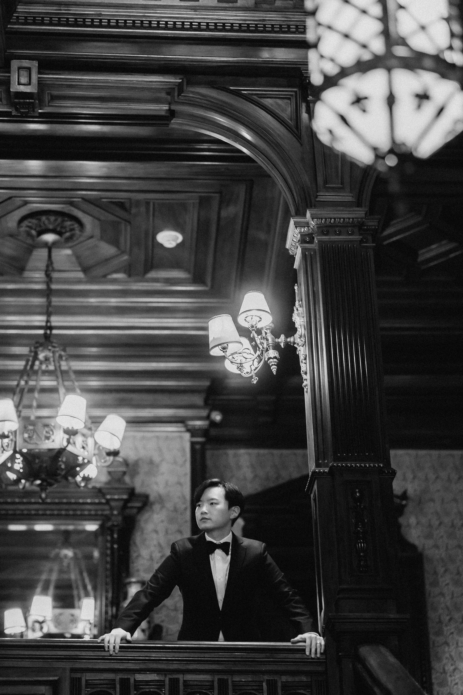 Black and white photo of groom on elegant vintage staircase with chandelier lighting.